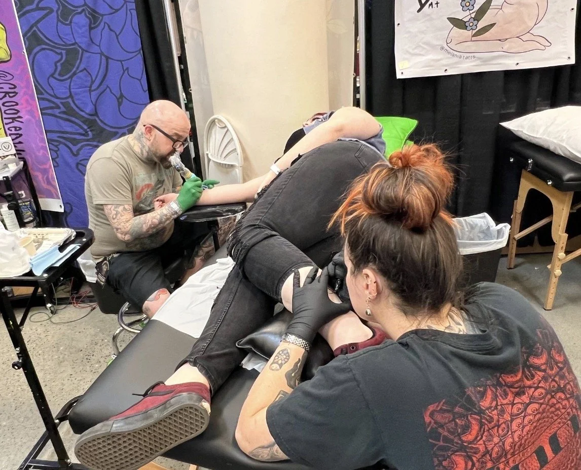 A woman with red hair getting a tattoo on her leg at a tattoo convention while a tattoo artist with glasses, tattooed arms, and gloves works on her tattoo. Another person, with tattoos on their arm, is leaning over to observe.
