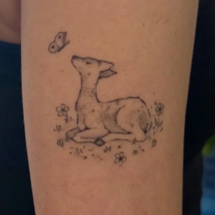 Tattoo of a sitting fawn with a butterfly above its head, surrounded by small flowers and grass.