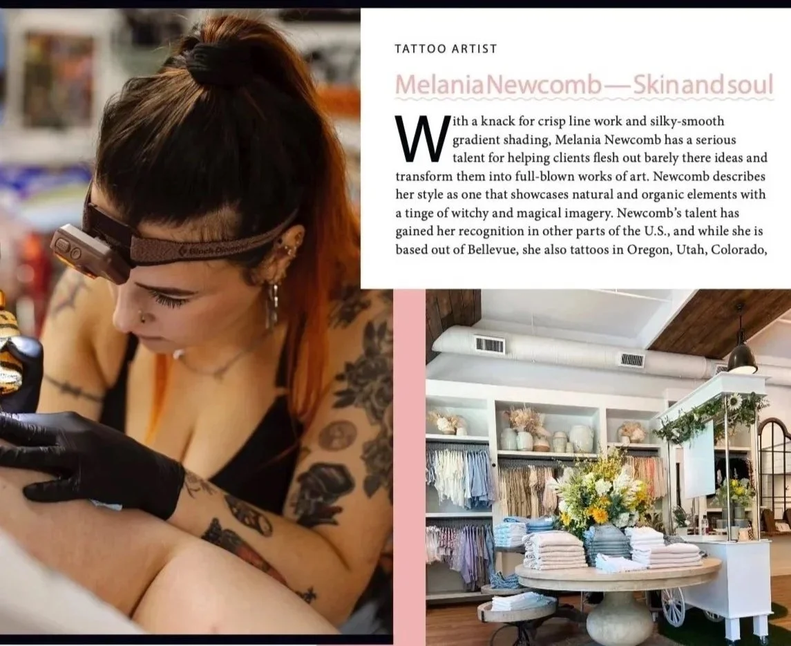 A tattoo artist working on a client's tattoo, with shelves of fabric or clothing and a table with a floral arrangement in a store interior.