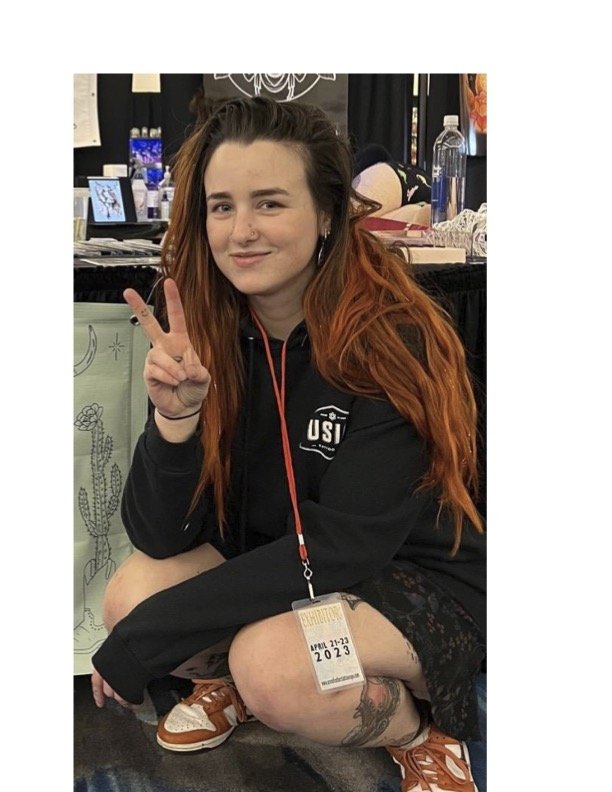 Young woman with long red hair squatting and making a peace sign, wearing a black hoodie, lace shorts, and sneakers, at an event with lanyard, badge, and various items in the background.