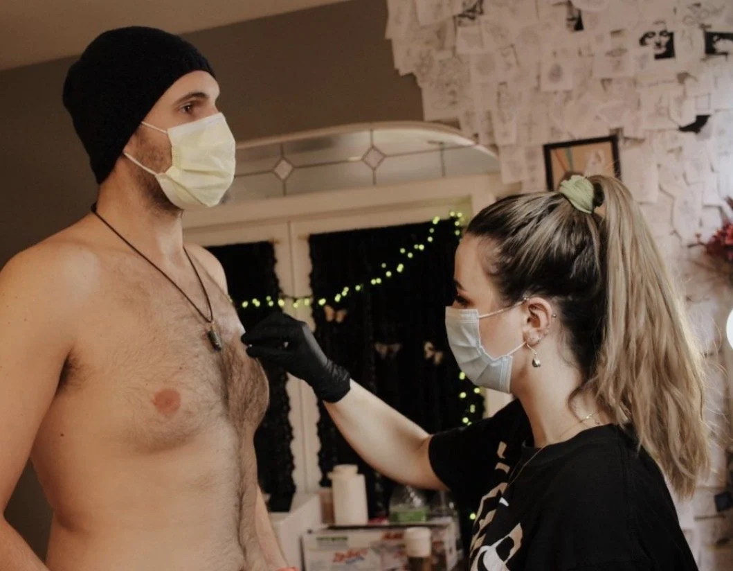 A woman in black scrubs, wearing a face mask and black gloves, is tattooing a shirtless man with a beanie and a face mask in a room decorated with string lights and artwork.