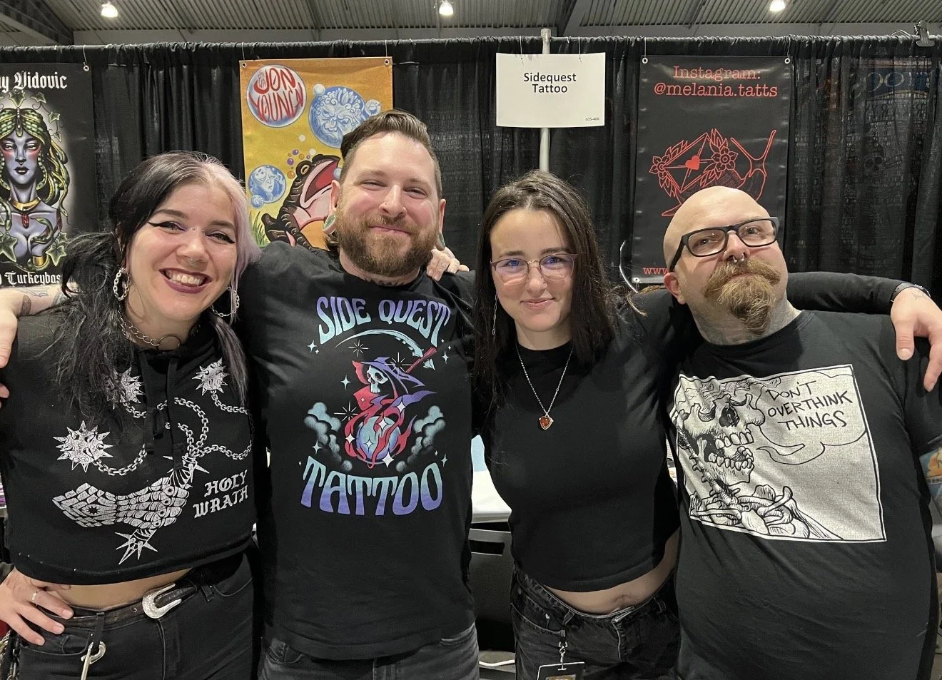 Four people standing at a tattoo convention, smiling with arms around each other, black booths with tattoo art and signs behind them.