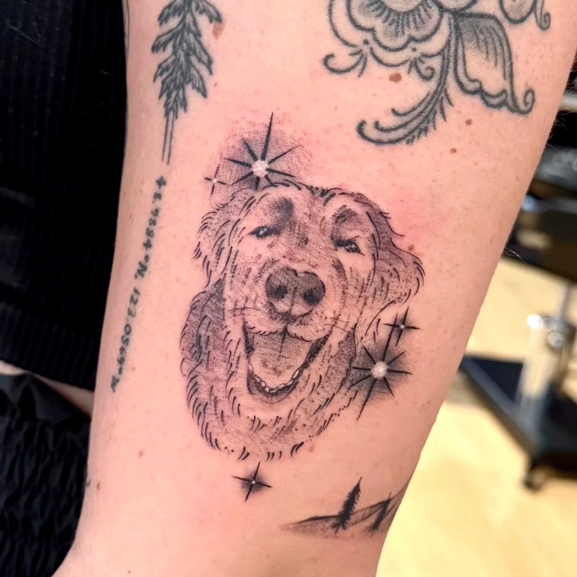 Close-up of a tattoo on skin, featuring a detailed illustration of a dog's face and various decorative elements including a star, words, and floral patterns.