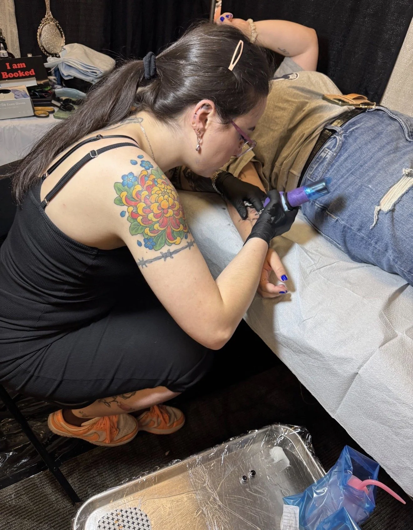 A woman with tattoos on her arm and dark hair tied back, wearing glasses and black clothing, is lying on a table receiving a tattoo on her forearm. She is wearing black gloves and using a tattoo machine. The scene appears to be in a tattoo parlor wit
