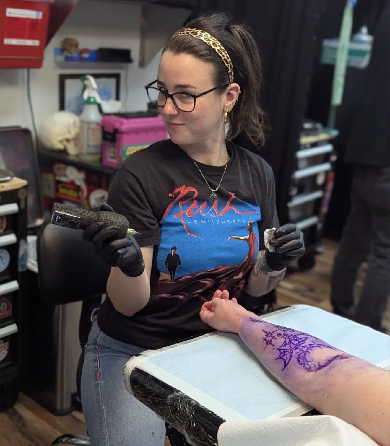 A woman with glasses and a leopard print headband tattoos a person's arm with purple ink at a tattoo shop. The person has a large tattoo on their forearm. The woman wears a black Rush band T-shirt and black gloves.