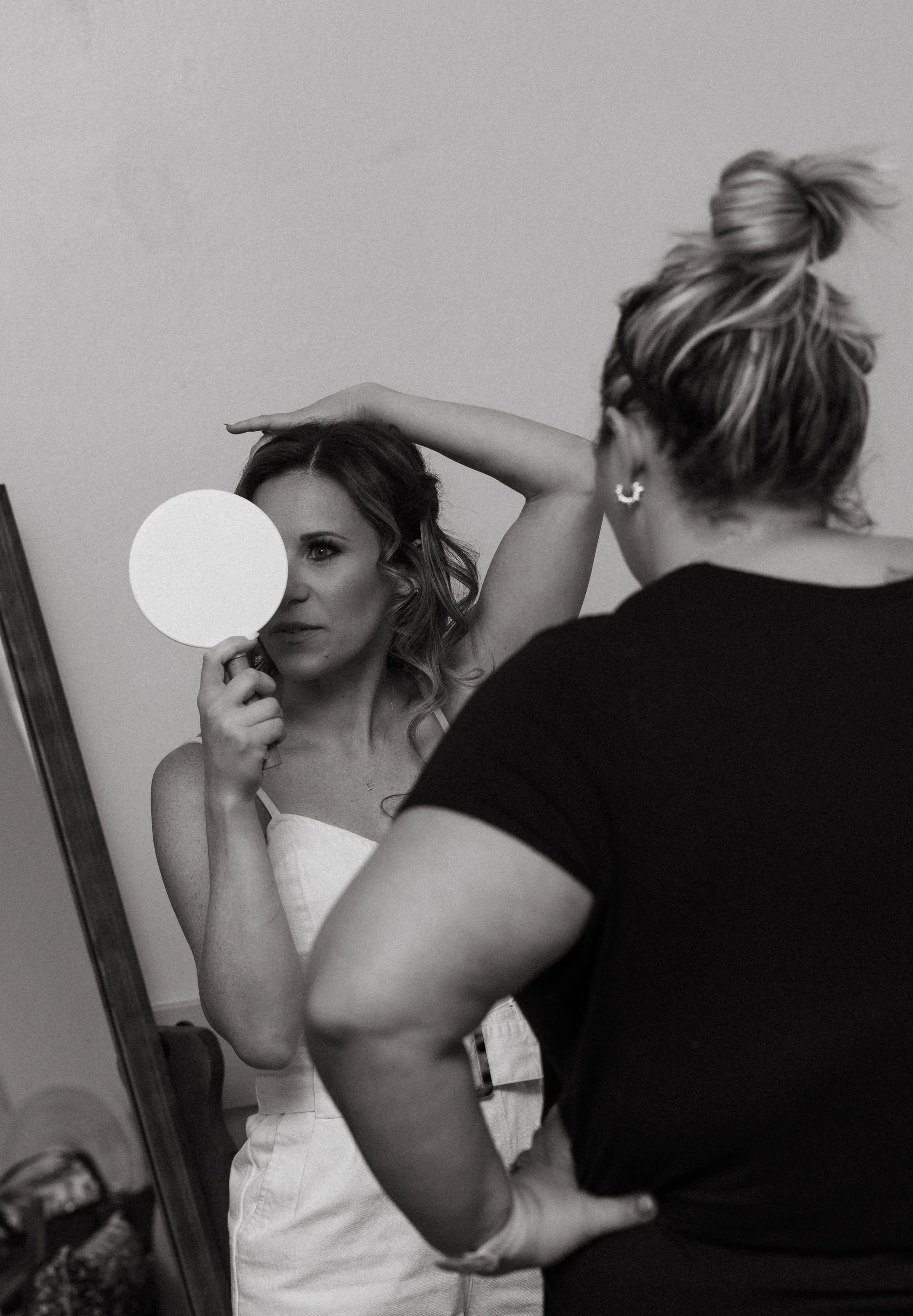 A woman looking in a mirror holding a round mirror in front of her face, while another woman, possibly a hairstylist, observes her from behind. The woman in the mirror has curled hair and wears a sleeveless top, and the hairstylist has her hair in a 