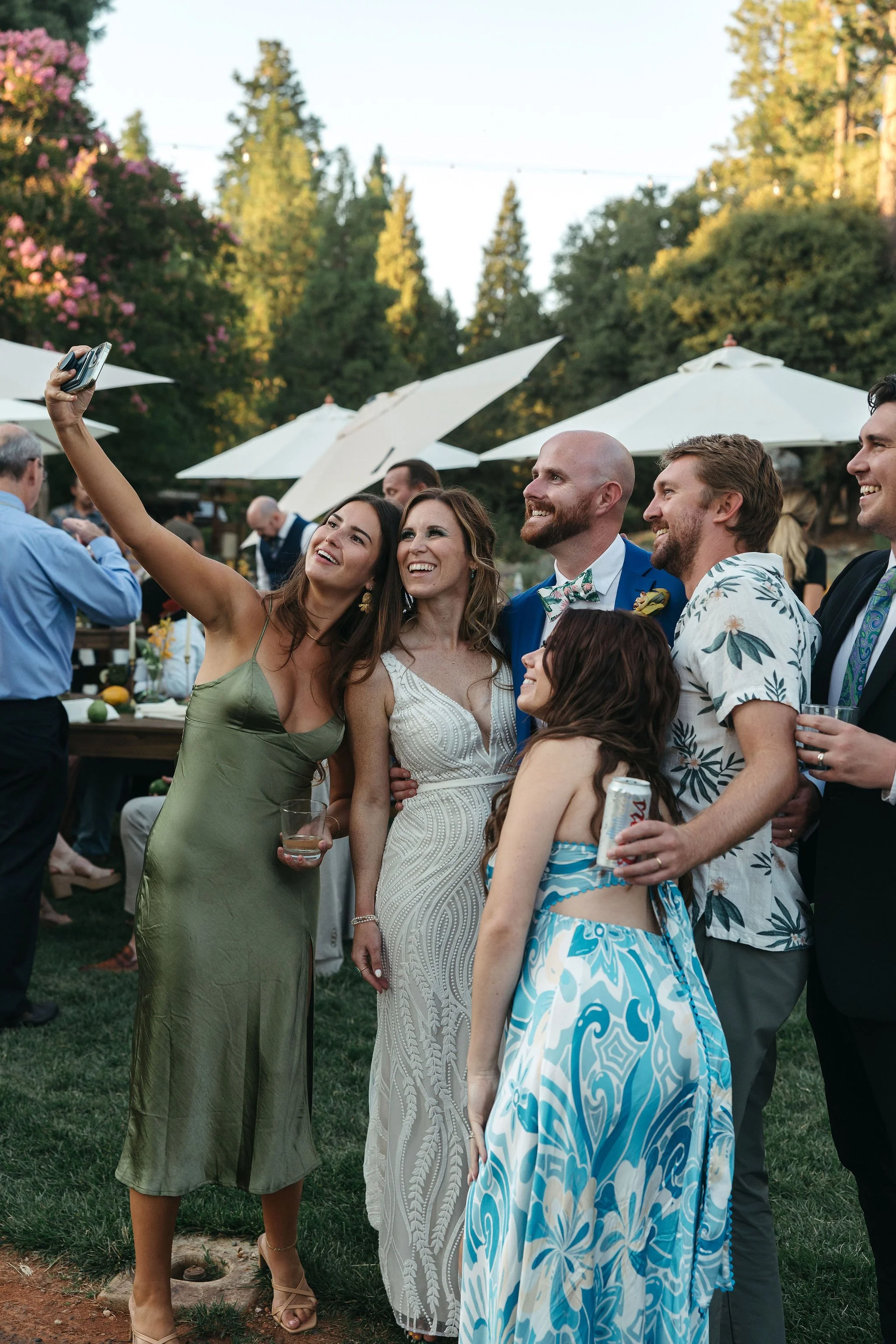 Group of people taking a selfie at an outdoor wedding reception, with wedding attire and decorations.