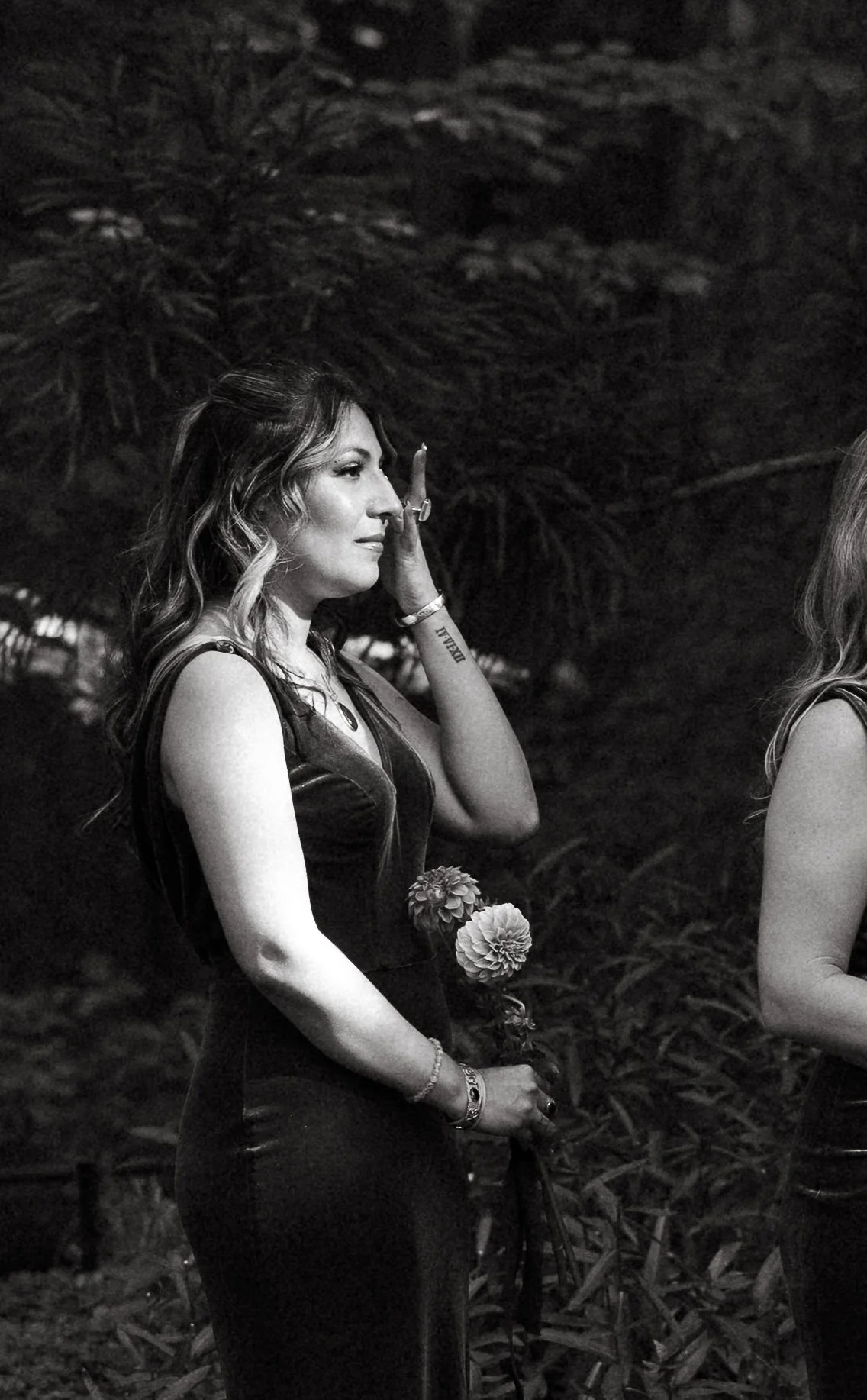 Black-and-white photo of a woman standing outdoors, holding flowers, with her hand touching her face, and looking contemplative.
