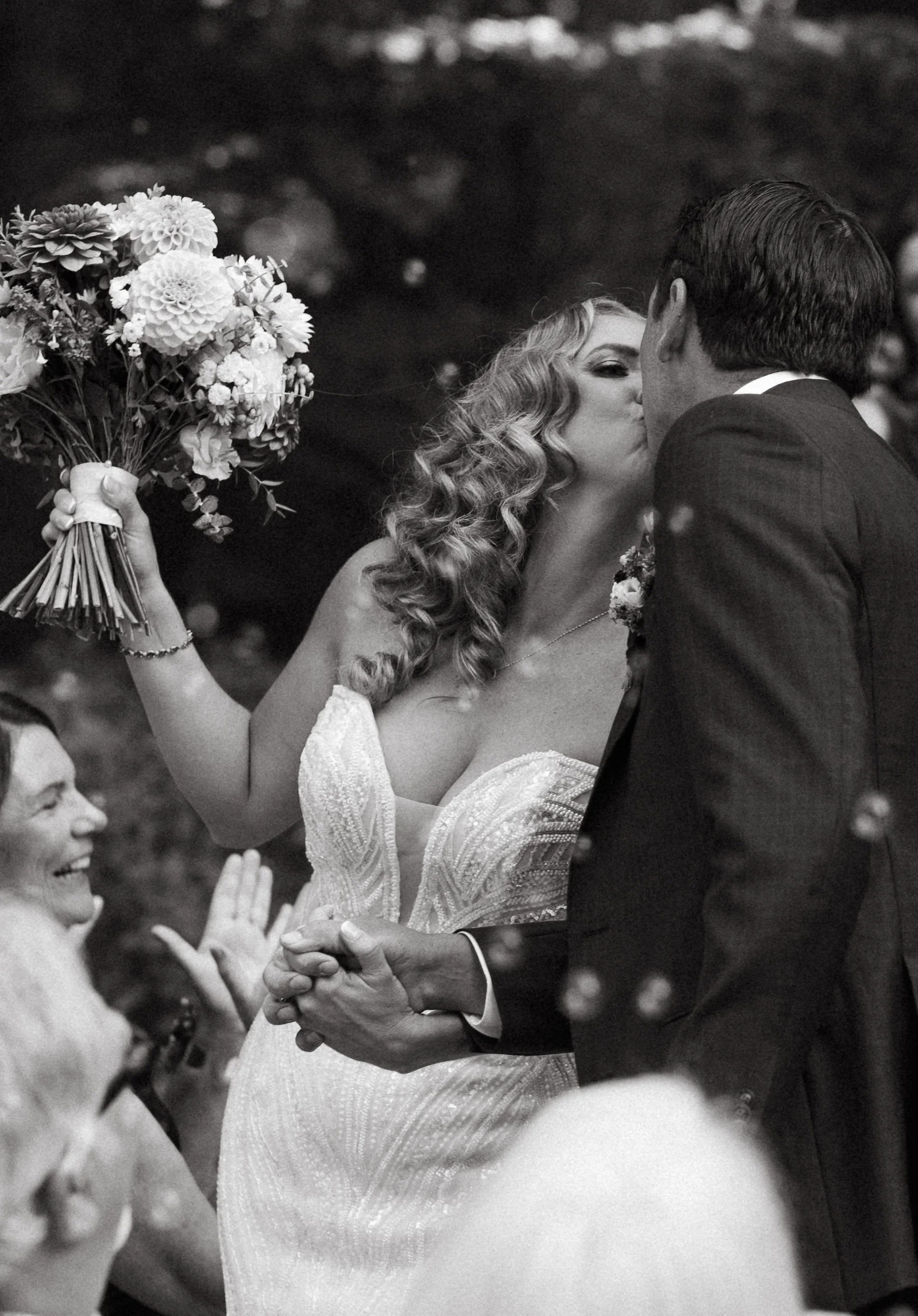 A bride and groom kissing during their wedding ceremony, with the bride holding a bouquet of flowers in her left hand and smiling woman witnessing the moment.