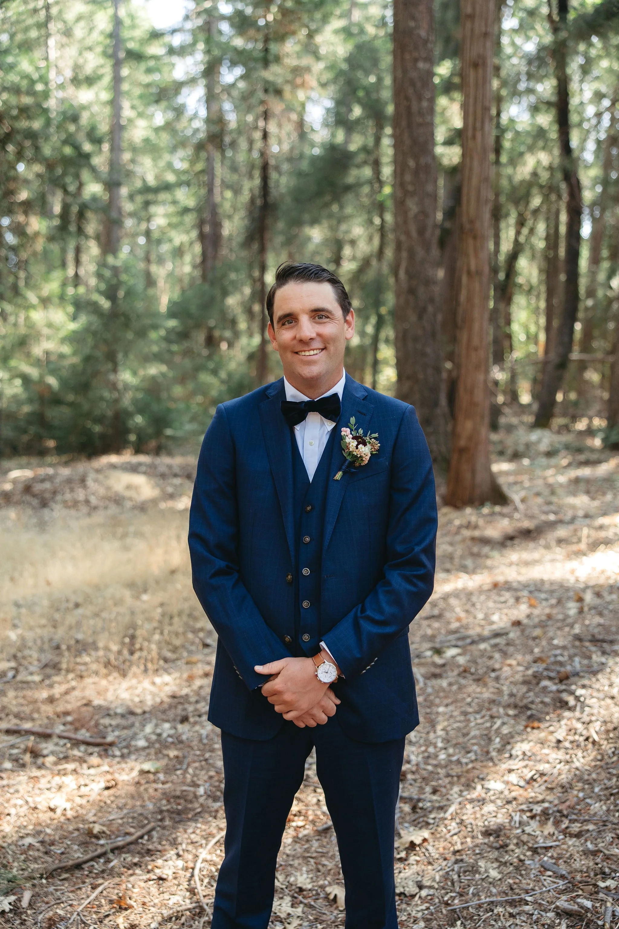 A groom dressed in a blue suit with a black bow tie and a boutonnière, standing outdoors in a forest with tall trees and sunlight filtering through the leaves.