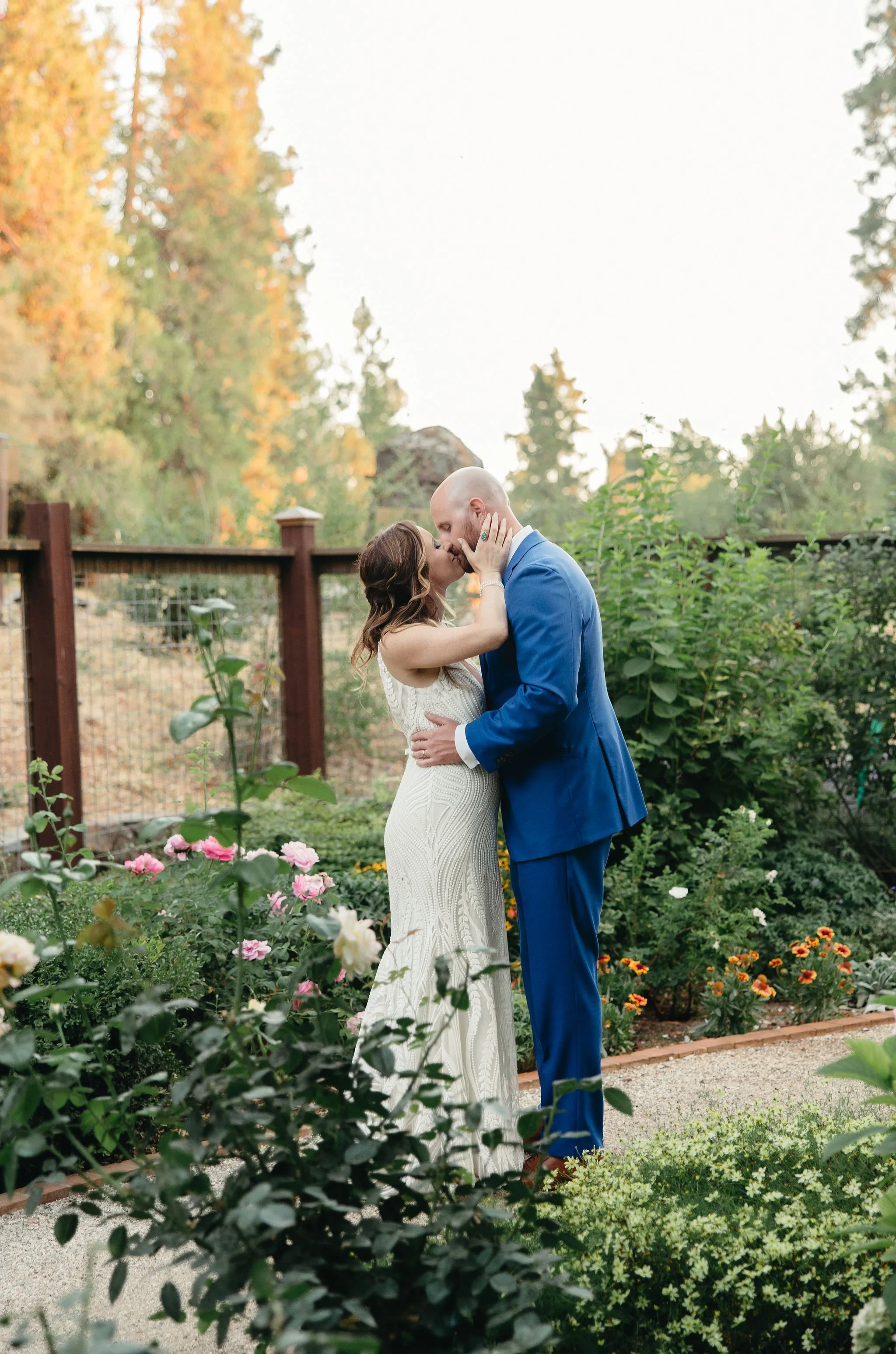A bride and groom kiss in a garden with flowers and greenery, the bride wearing a white dress and the groom in a blue suit, during sunset.