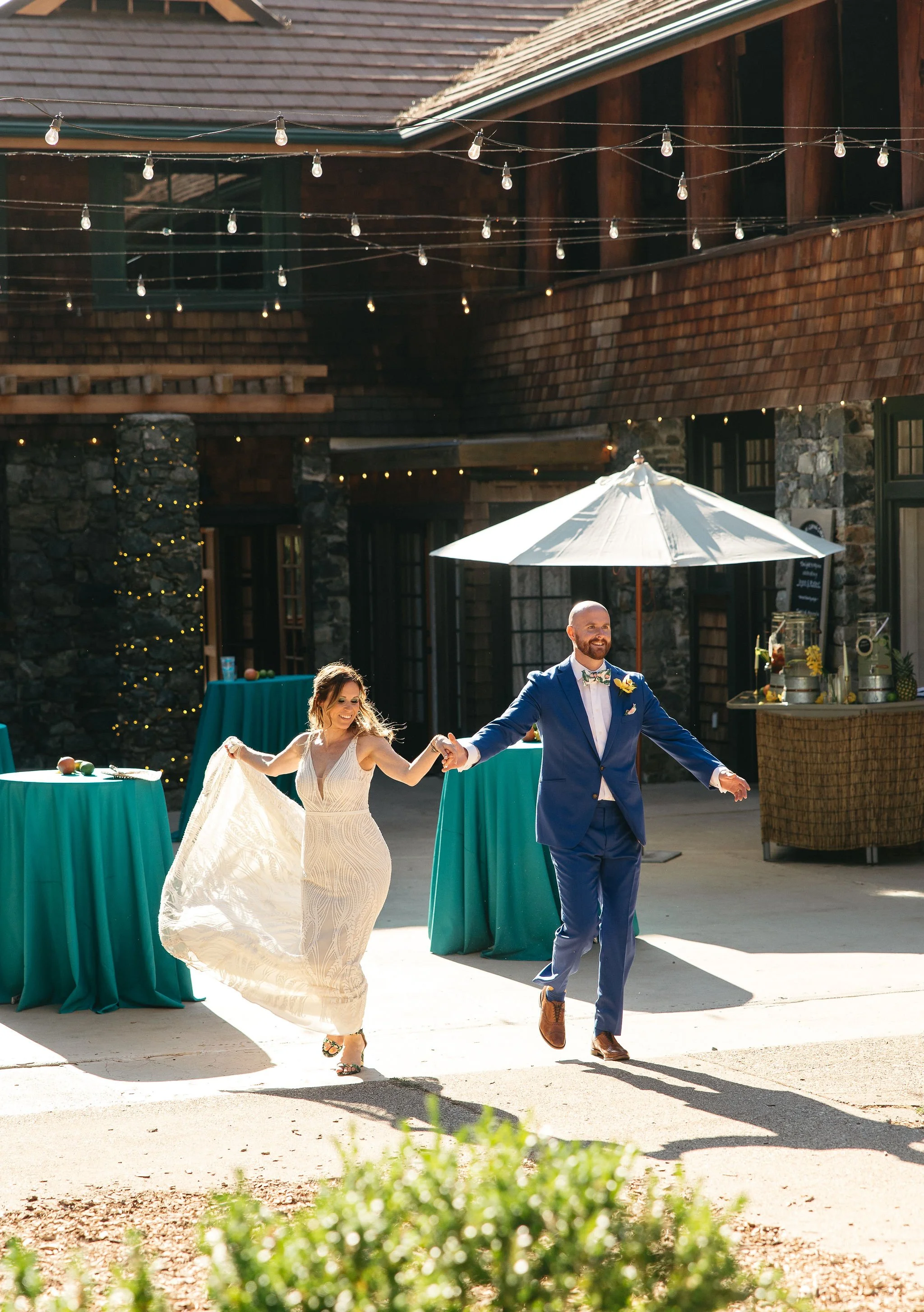 A newlywed couple dancing outdoors under string lights and a white patio umbrella in a rustic venue with stone walls and wooden roof.