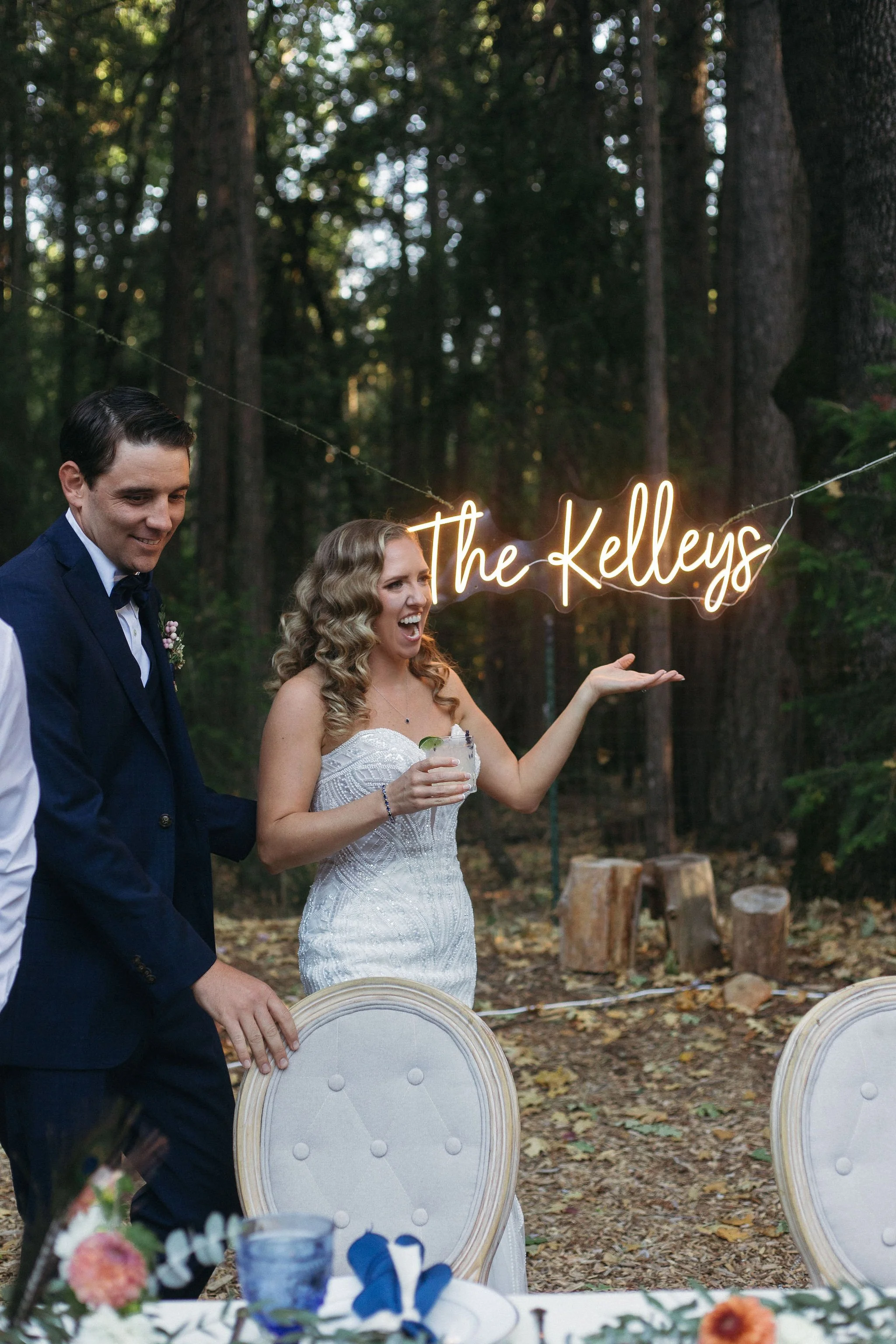 A wedding celebration outdoors with a woman in a white dress and a man in a dark suit. The woman is holding a drink and gesturing towards a neon sign that reads 'The Kelleys' hanging in a wooded area.