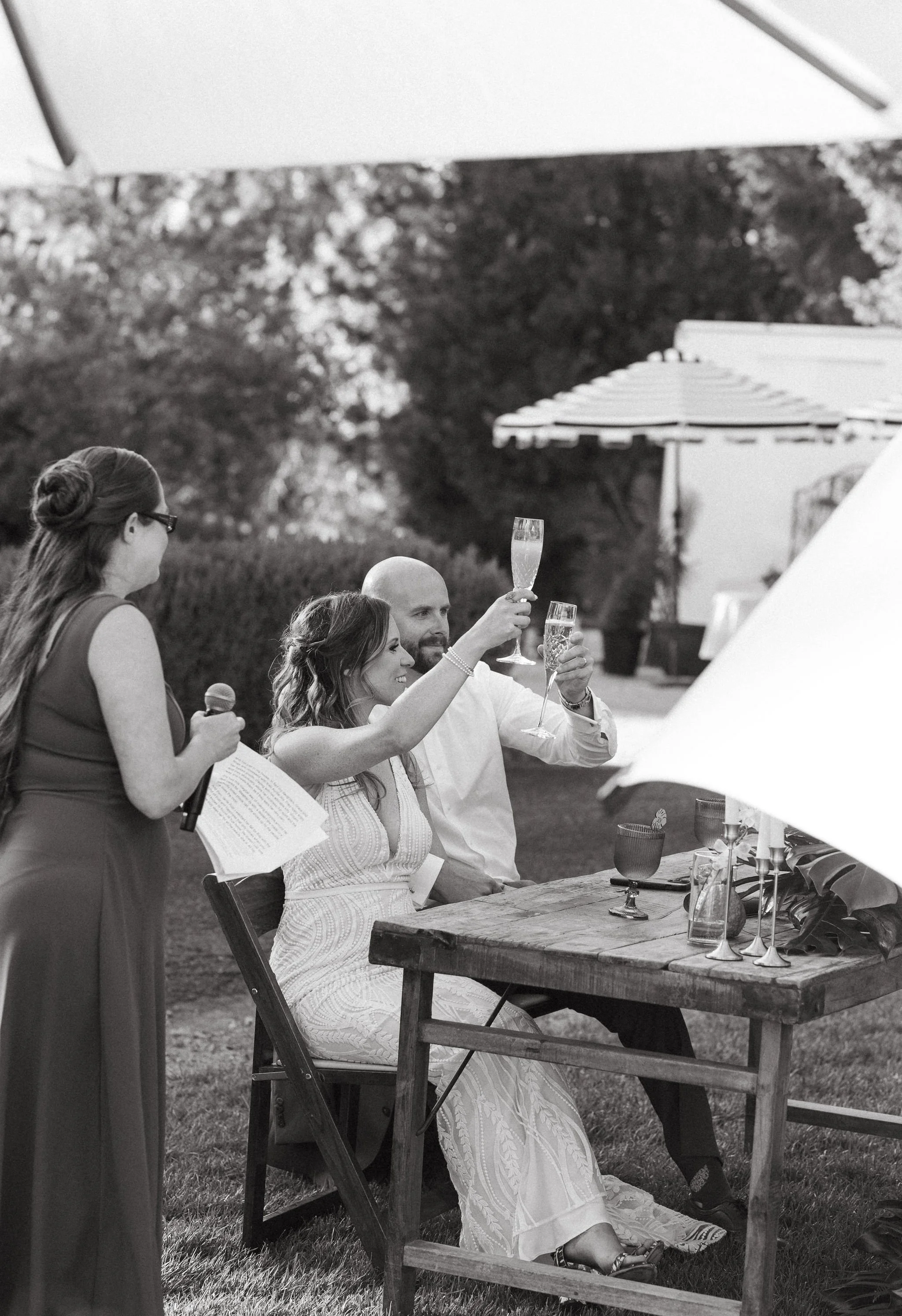 A black and white photo of a couple sitting at a rustic wooden table outdoors, raising glasses in a toast, with a woman standing nearby holding a microphone and papers. The scene is set on a lawn with trees and umbrellas in the background.