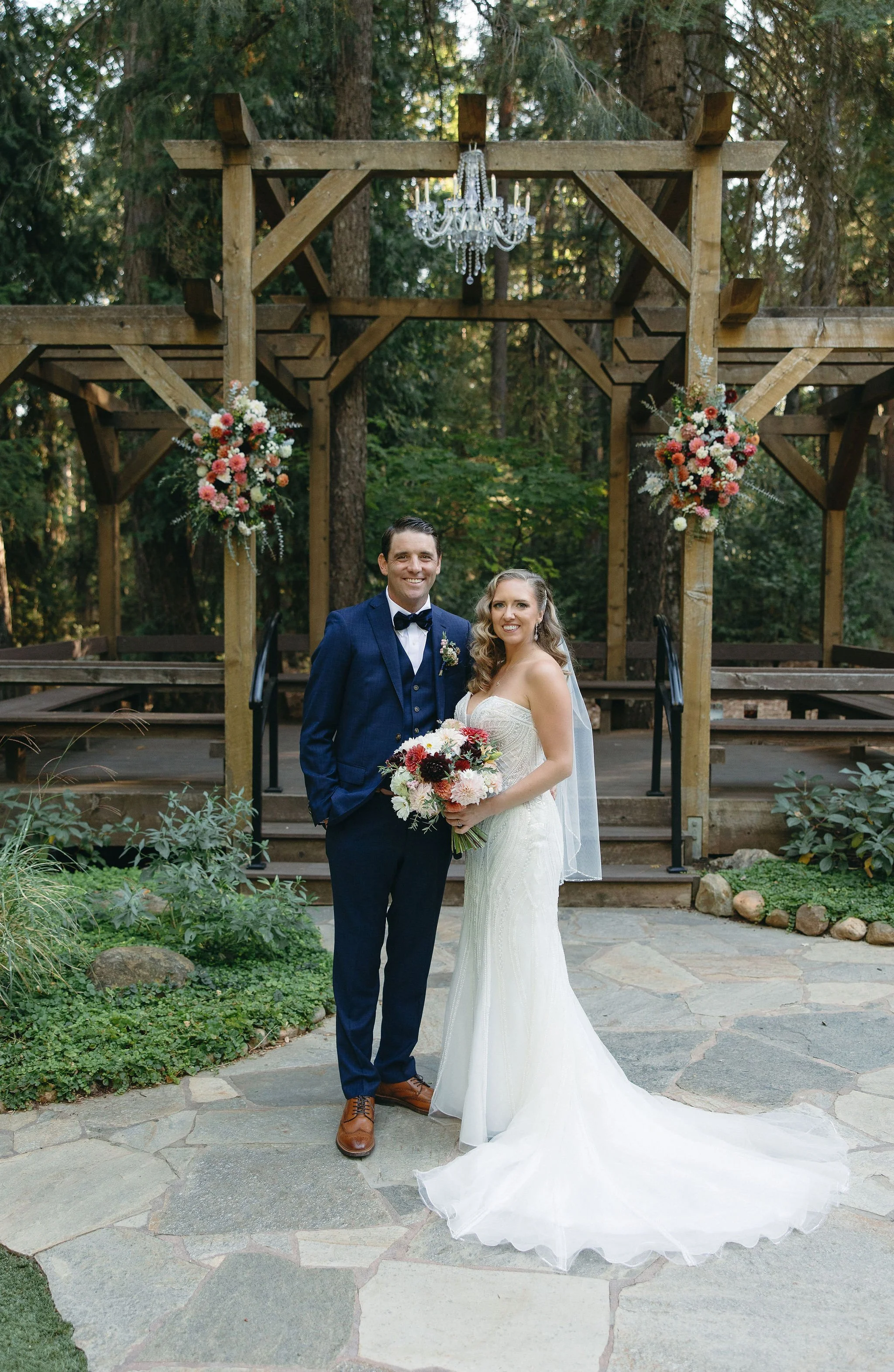 A bride and groom pose together outdoors in front of a wooden wedding arch decorated with flowers and a chandelier. The bride wears a white wedding gown and holds a bouquet, while the groom wears a navy suit with a bow tie.