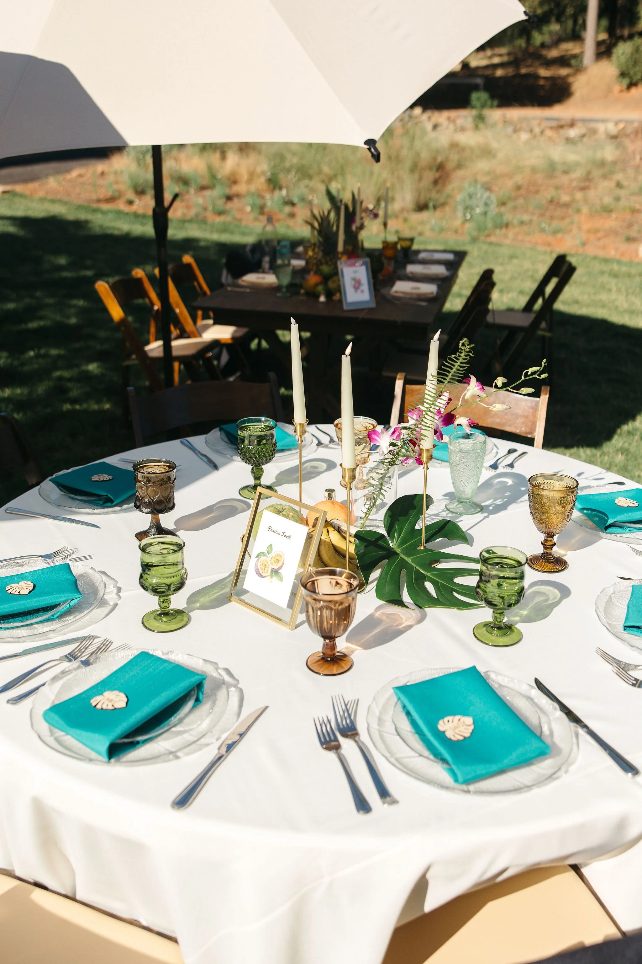 Round outdoor dining table set for a celebration with colorful glassware, turquoise napkins, and tropical-themed decor, shaded by a white umbrella in a garden.