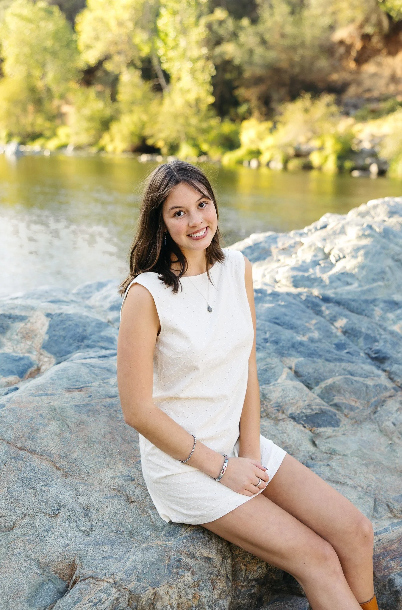 A young woman sits on large rocks by a river with trees in the background during daylight, smiling at the camera.