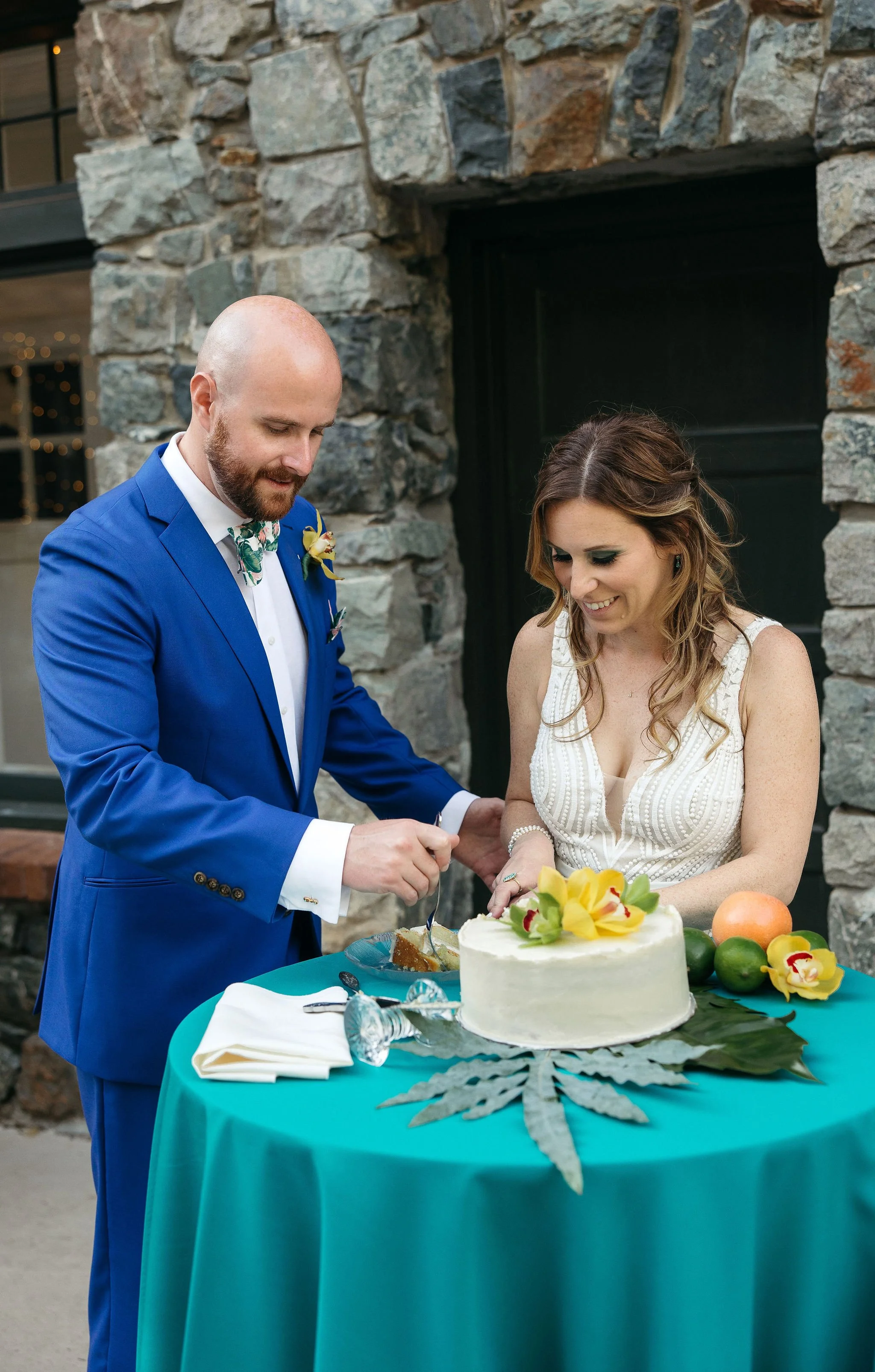 A bride and groom are cutting a wedding cake together. The groom is in a blue suit with a floral bowtie, and the bride is in a white dress. They are standing behind a teal table with a cake decorated with yellow flowers and fruit.
