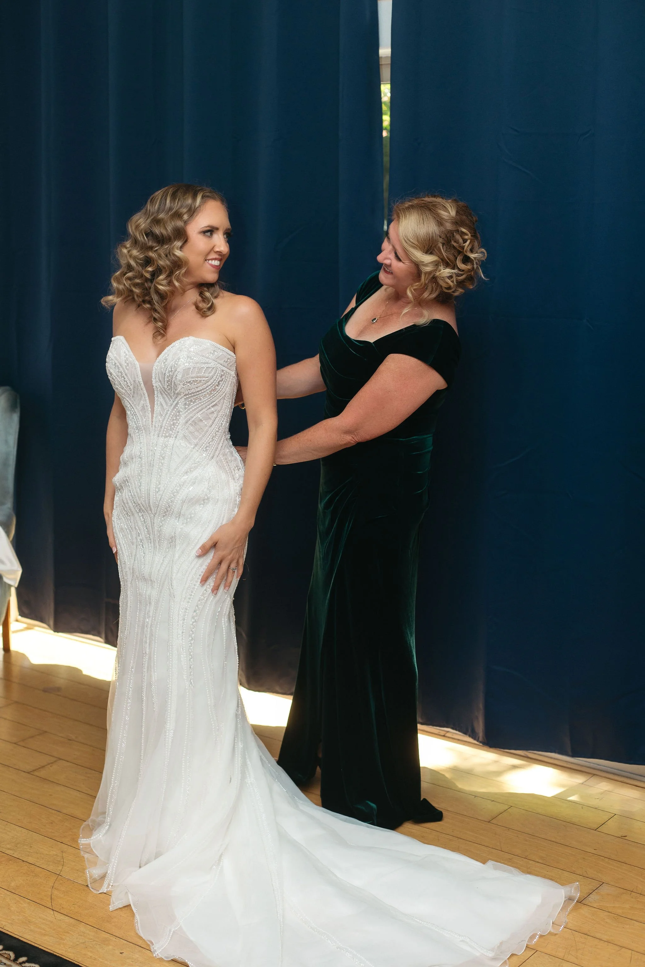 A woman in a white wedding dress getting assistance from another woman in a dark green dress, standing in front of blue curtains.