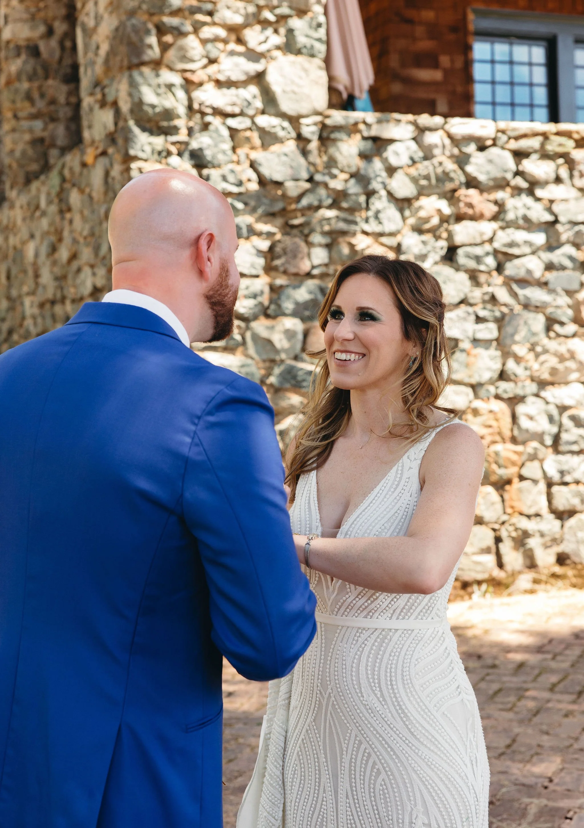 A woman in a white wedding dress smiling and holding hands with a man in a blue suit, in an outdoor setting with a stone wall background.