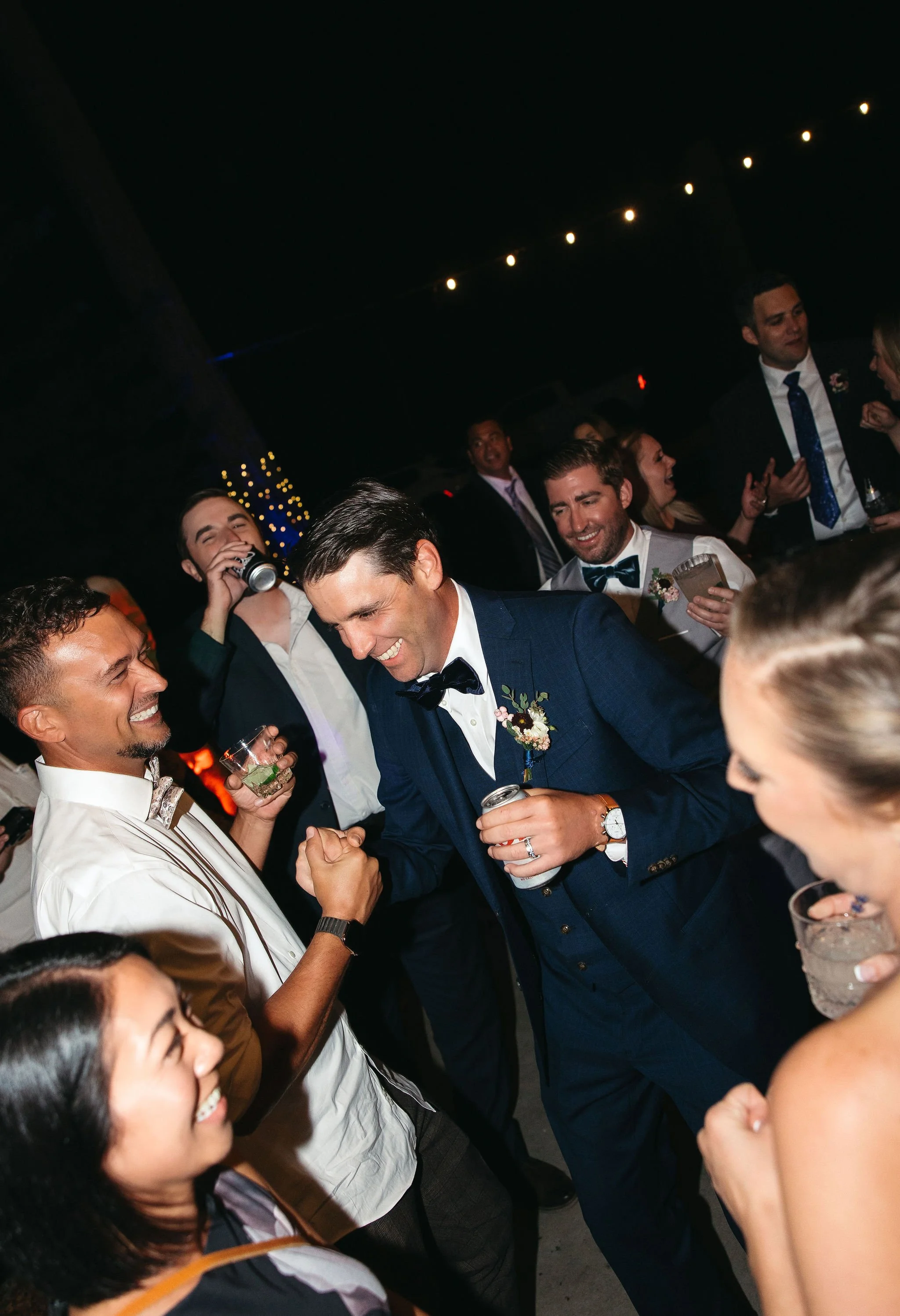 Group of people at a celebration, with two men in the center shaking hands and smiling, surrounded by smiling friends holding drinks under string lights at night.