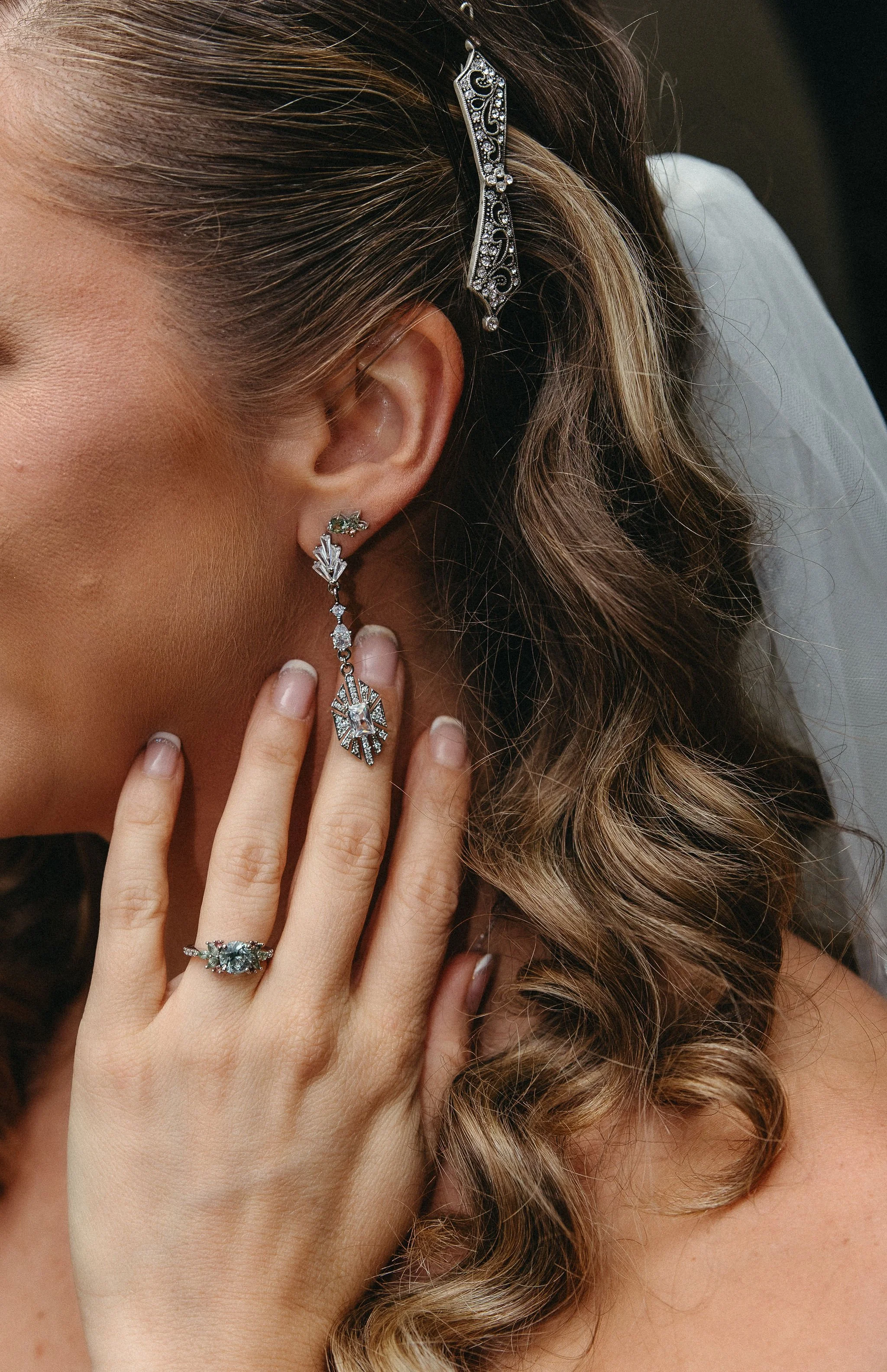 Close-up of a woman's face, showing elaborate jewelry including earrings and a hair clip, with curly hair and a pearl veil in the background.