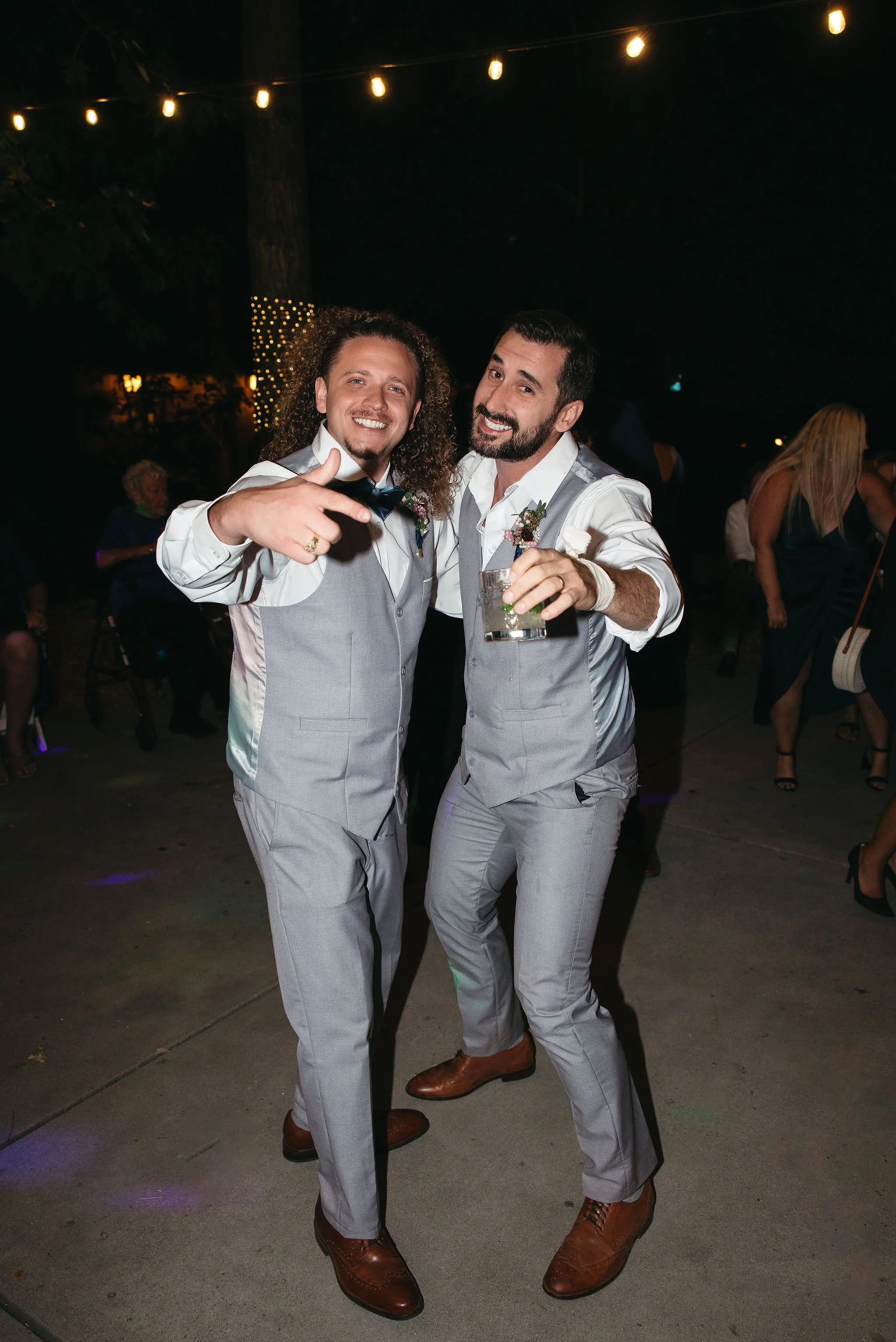 Two men dressed in light gray vests and pants at a nighttime event, smiling and posing for the camera, one pointing with his finger, the other holding a drink, with string lights overhead and people in the background.