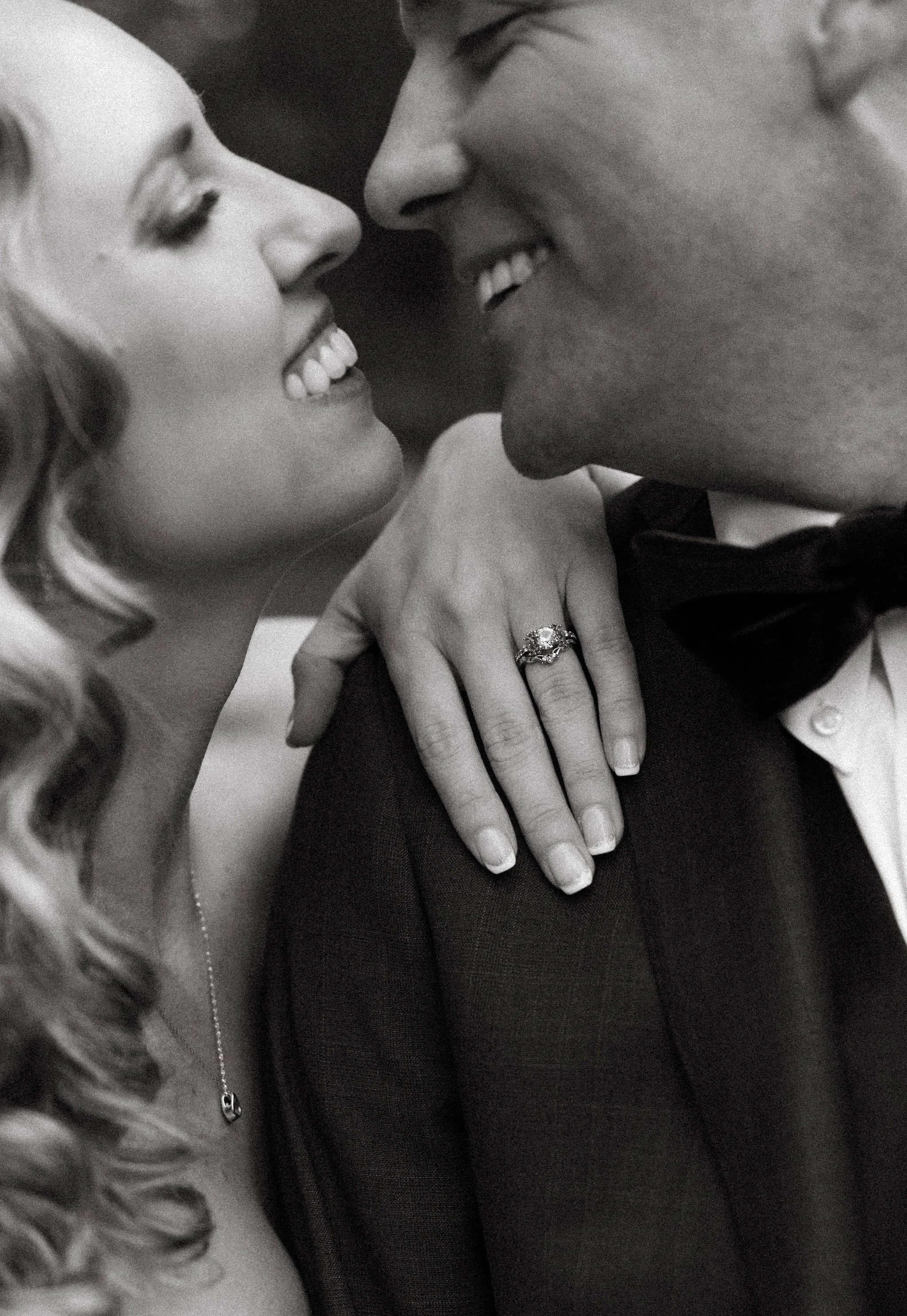 A black and white close-up of a couple smiling as they lean their foreheads together, with the woman's hand on the man's shoulder showing an engagement ring.