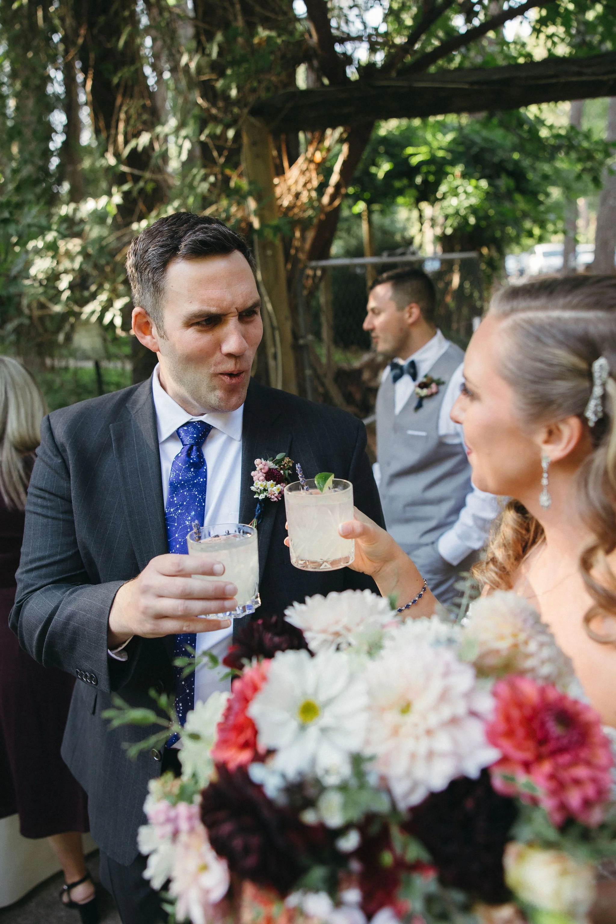 A bride and groom toasting with drinks at an outdoor wedding, with the bride holding a bouquet of flowers and a man in a suit in the background.