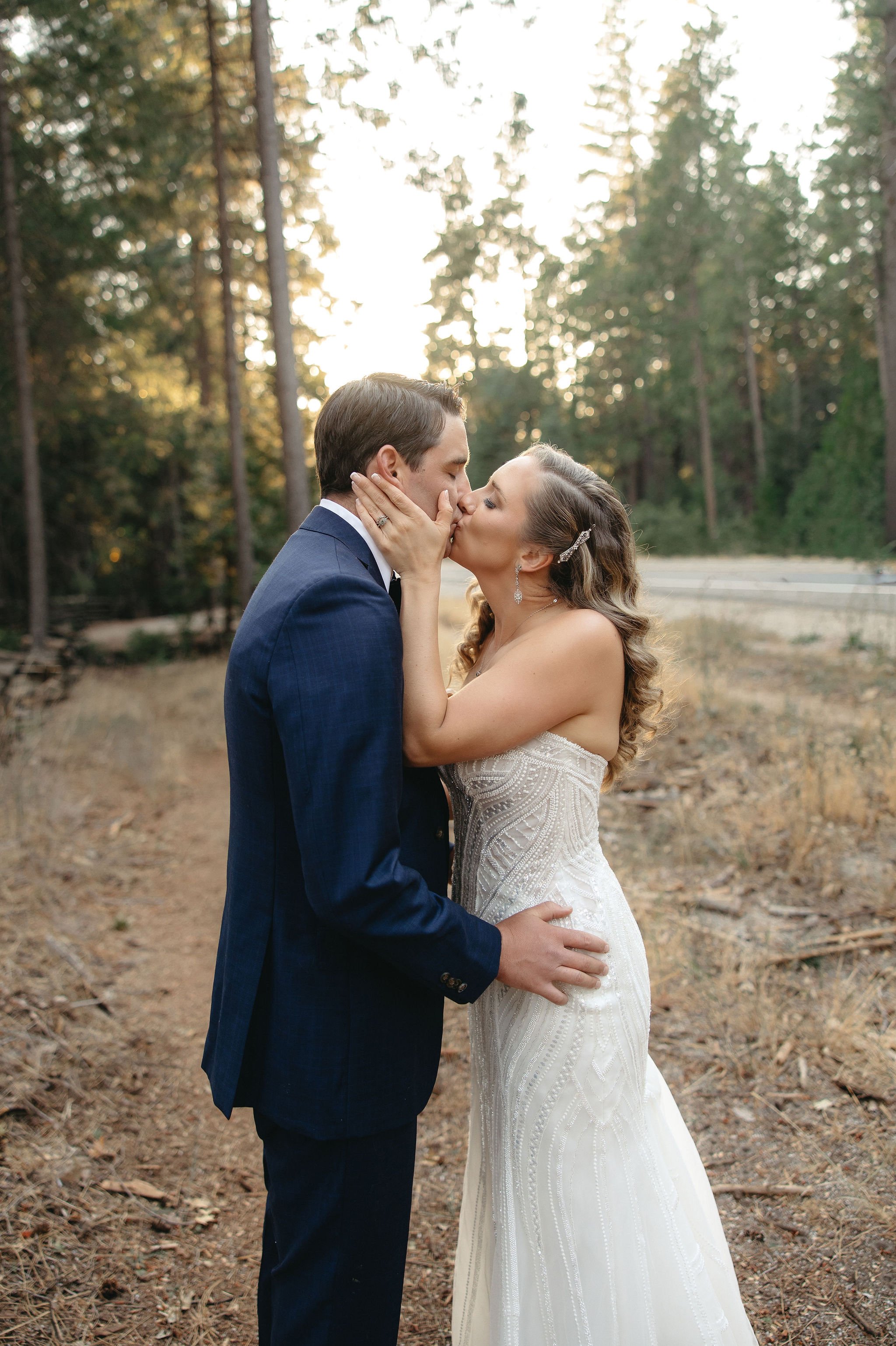 A couple in wedding attire sharing a kiss in a wooded outdoor setting during sunset