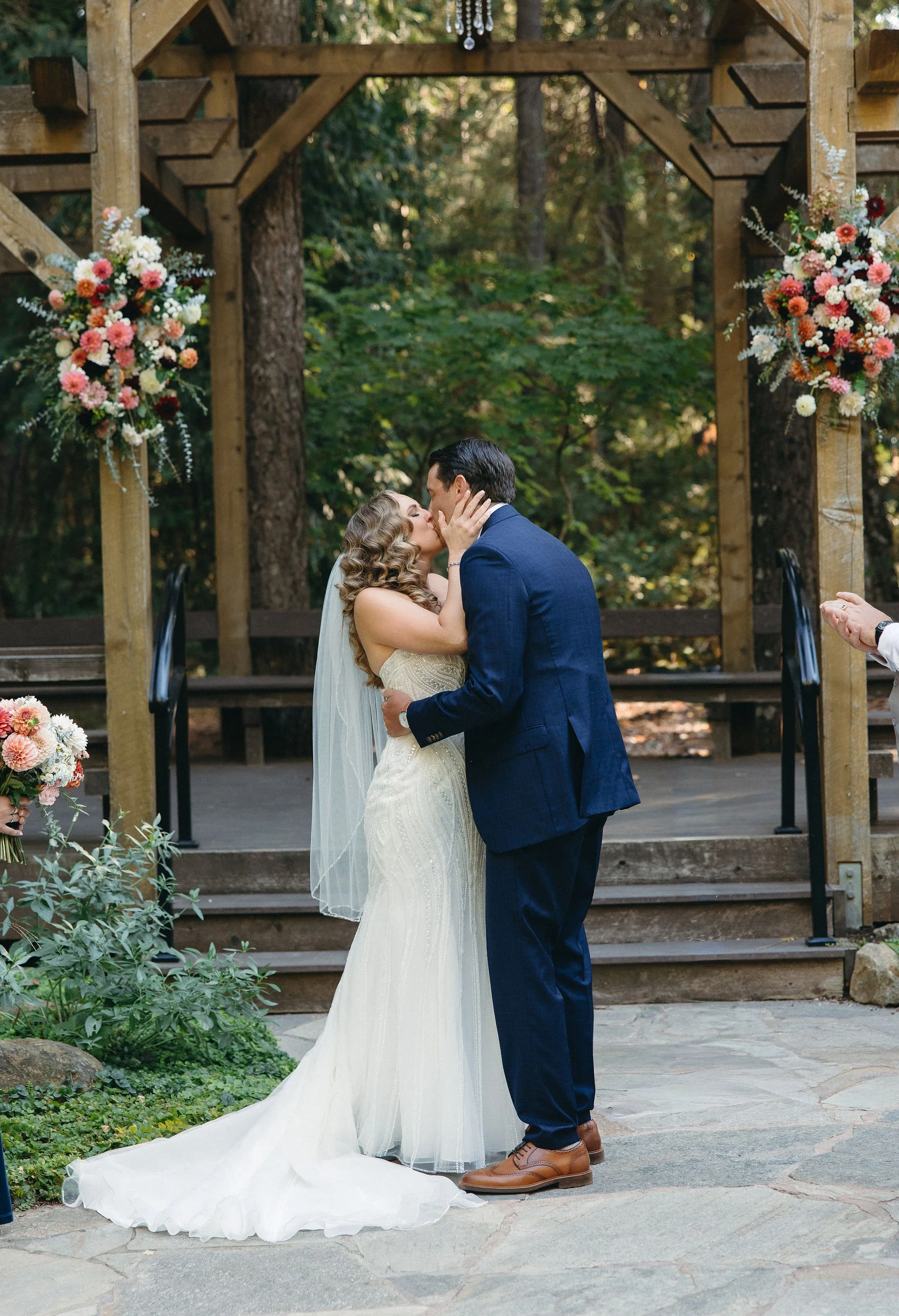 A bride and groom kiss during their outdoor wedding ceremony under a wooden arch decorated with colorful flowers, in a forest setting.