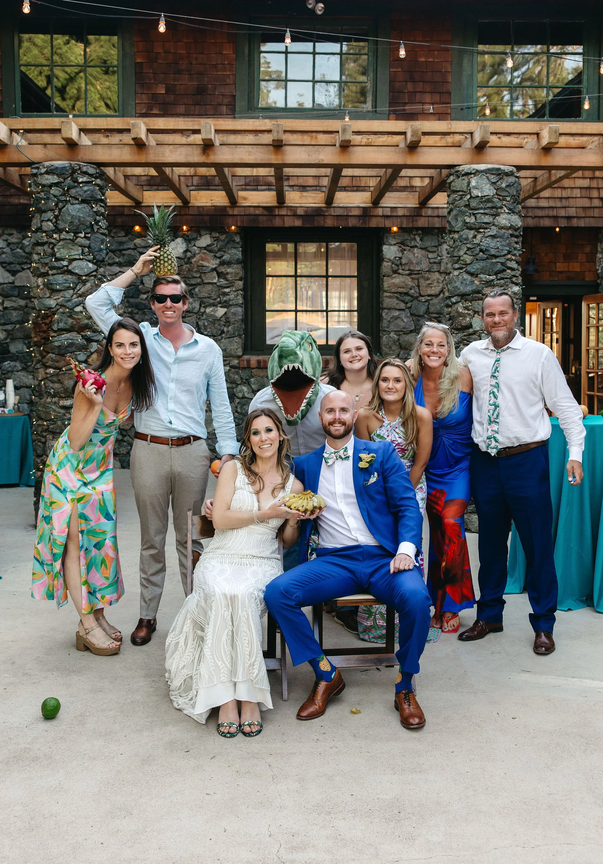 Group of friends in colorful attire celebrating outdoors with a rocky brick building in the background, some holding props, one dressed as a dinosaur, with festive string lights overhead.