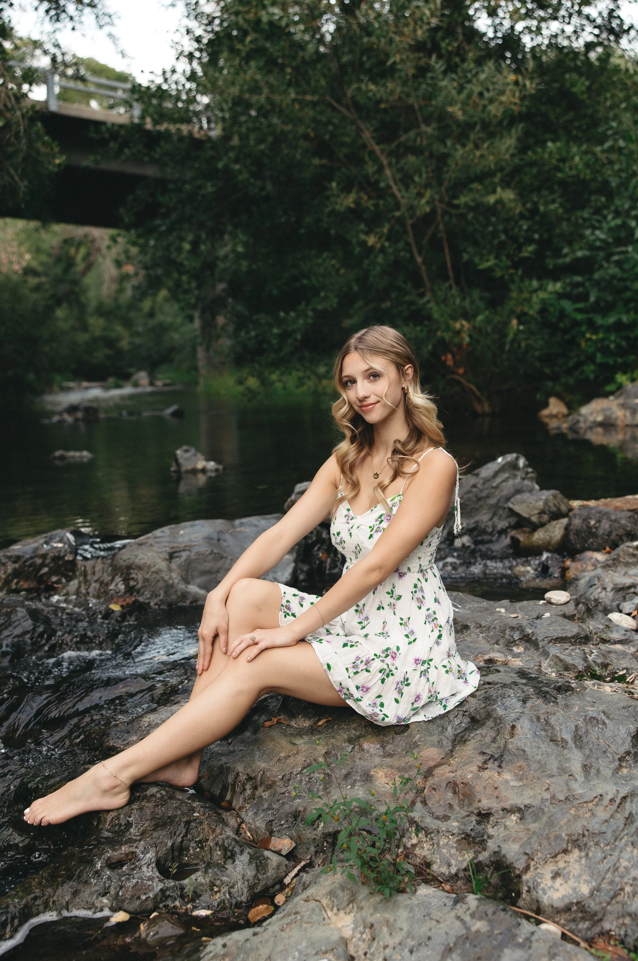 A young woman with long wavy blonde hair wearing a white dress with a green floral pattern sitting on rocks by a river in a lush green forest, with a bridge visible in the background.