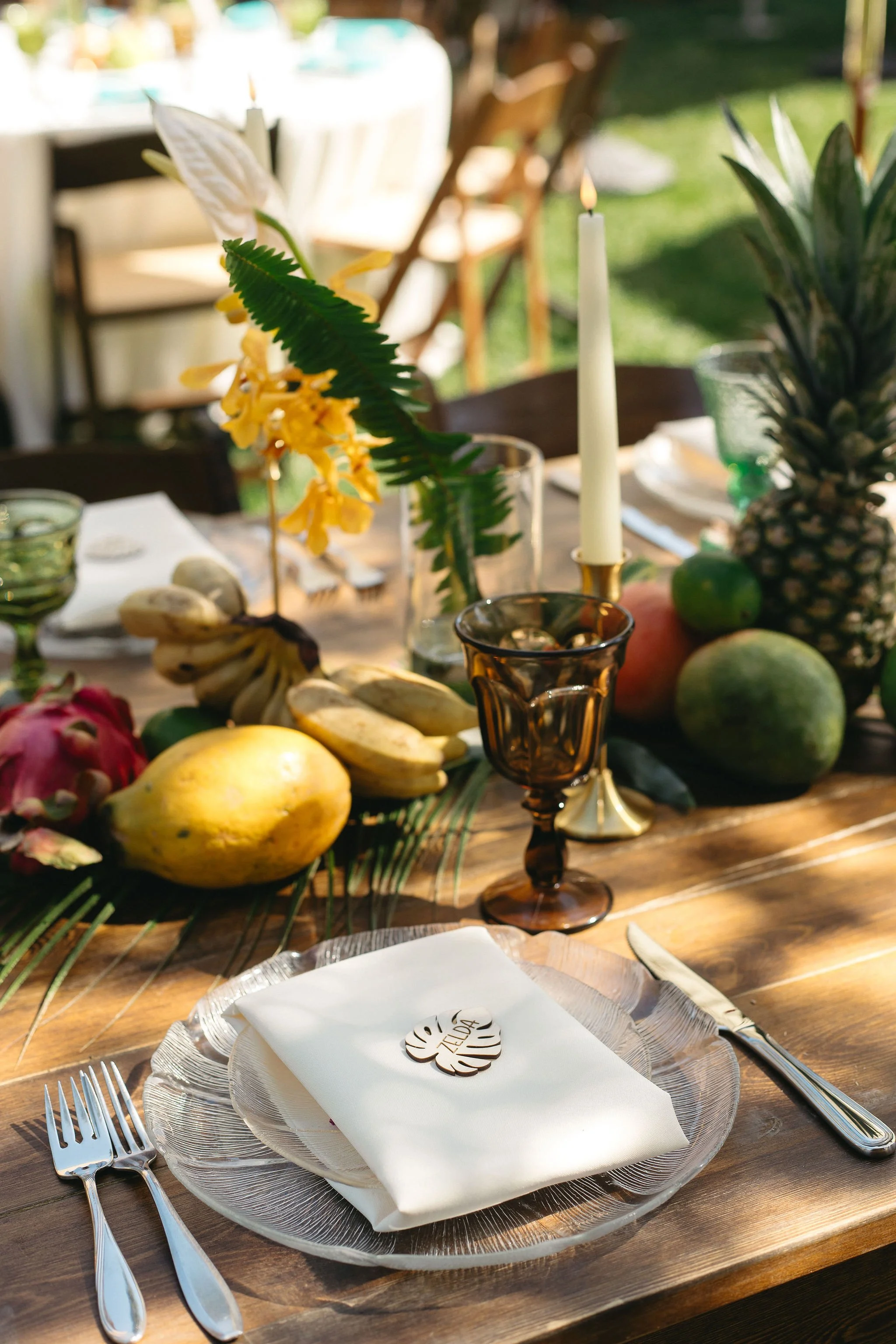 Tropical-themed table setting with a white napkin, a place card labeled 'ZARA', silverware, and a centerpiece of fruits, flowers, candles, and greenery.