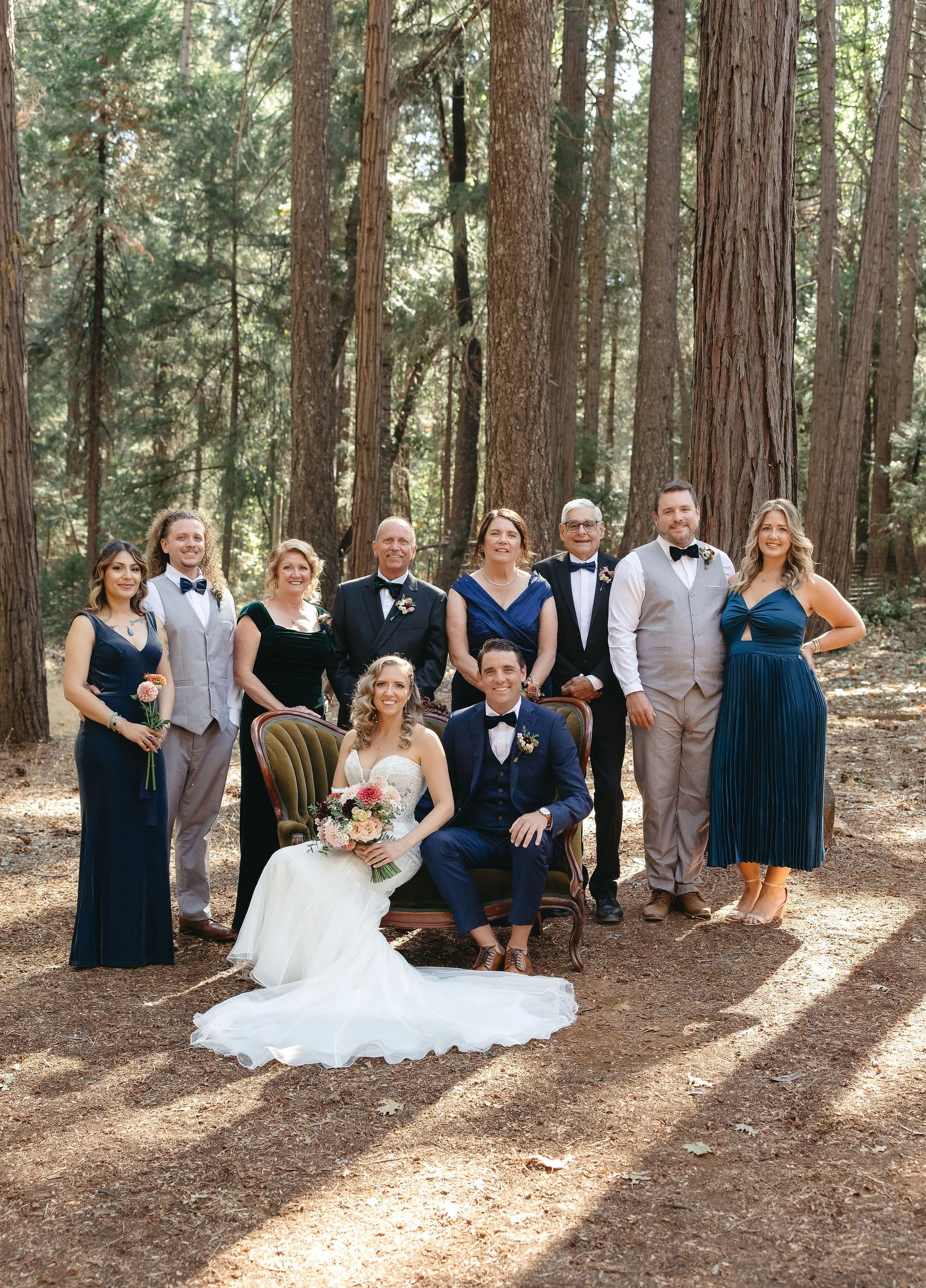 A group photo of a wedding party outdoors in a forest, with the bride and groom seated in the front and family and friends standing behind them.