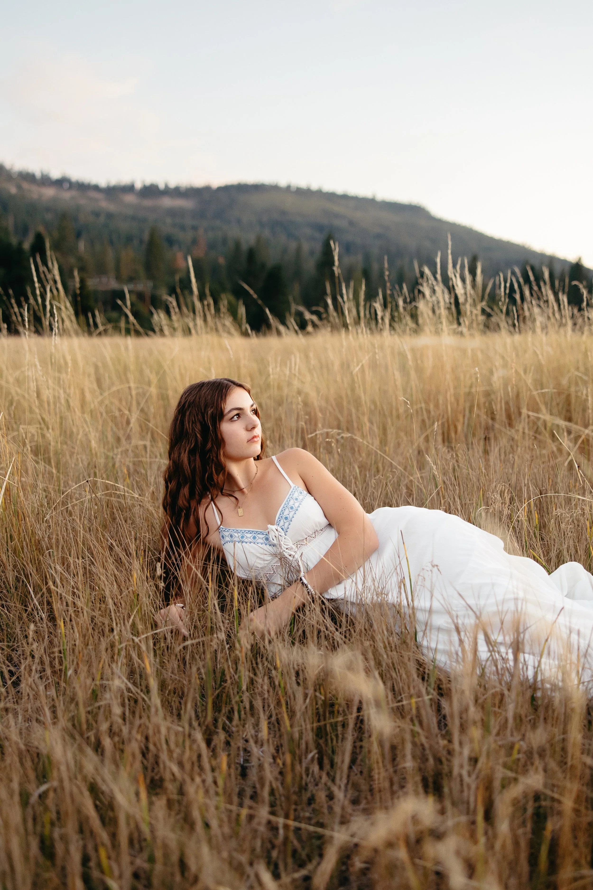 A young woman with long brown hair lying in a field of tall golden grass, with hills and trees in the background, during sunset.