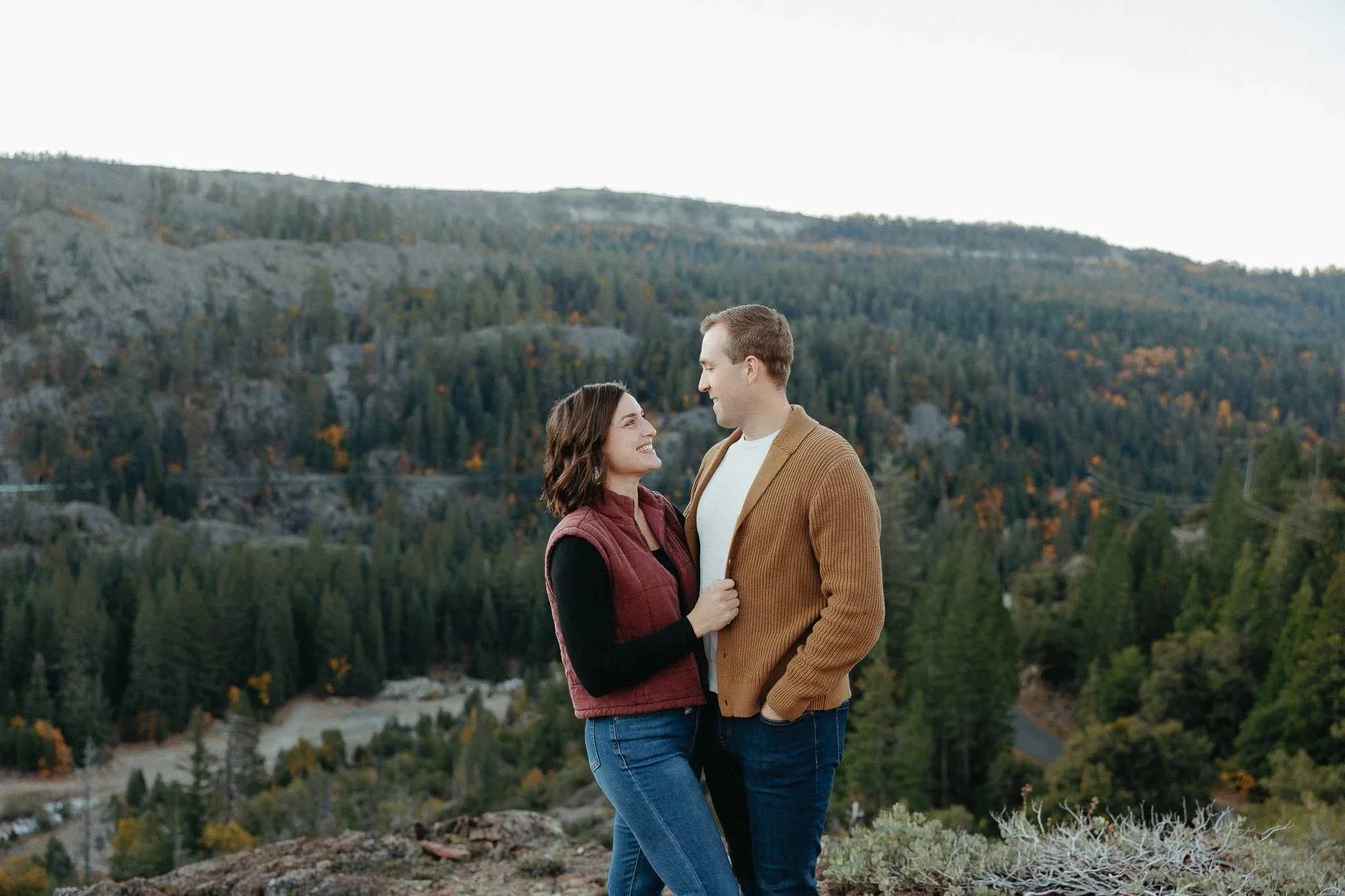 A smiling couple stands close together outdoors, facing each other with a scenic mountain and forest background during daytime.