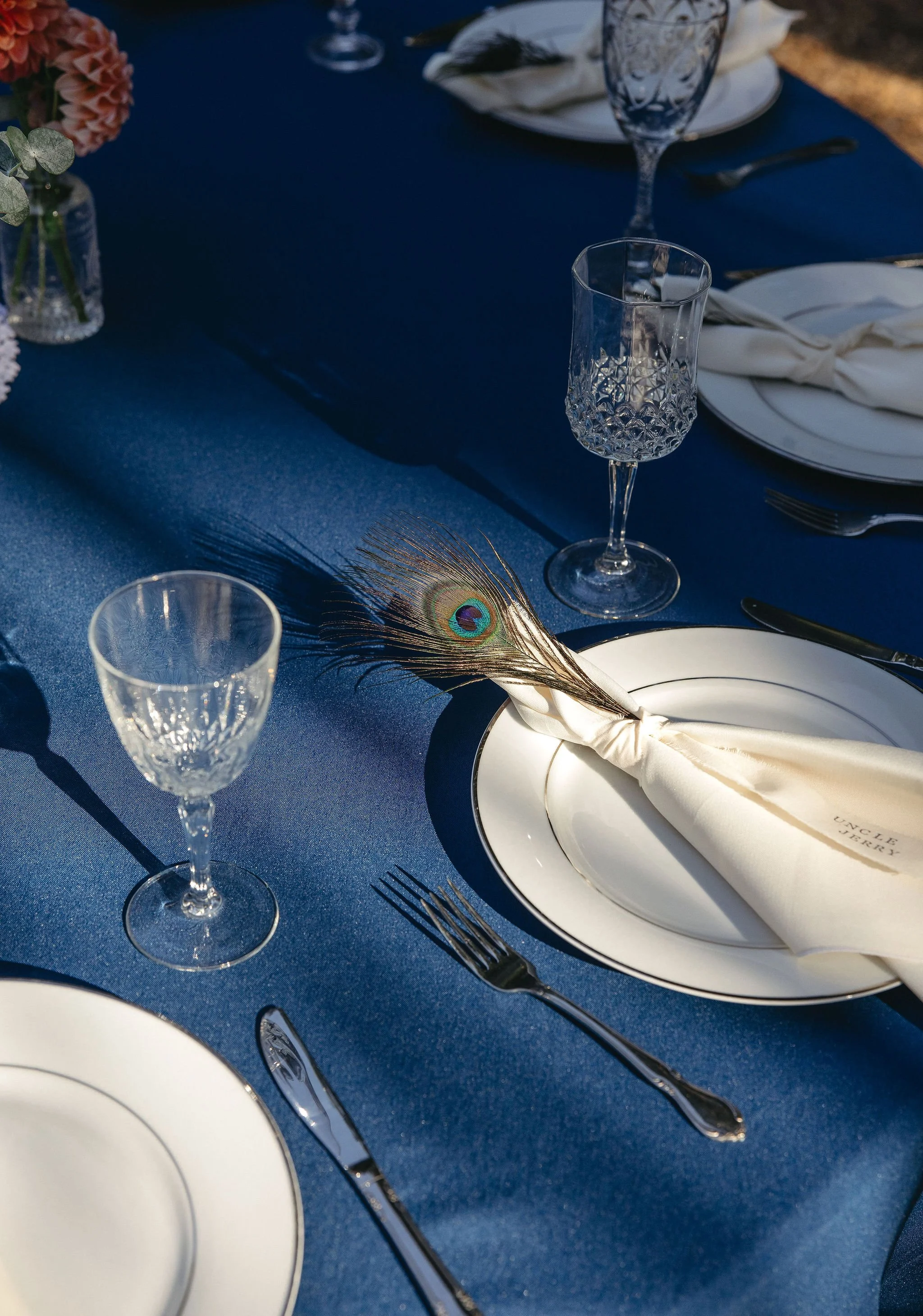 Elegant dining table set with white plates, crystal glasses, silverware, a navy blue tablecloth, and a decorative peacock feather.