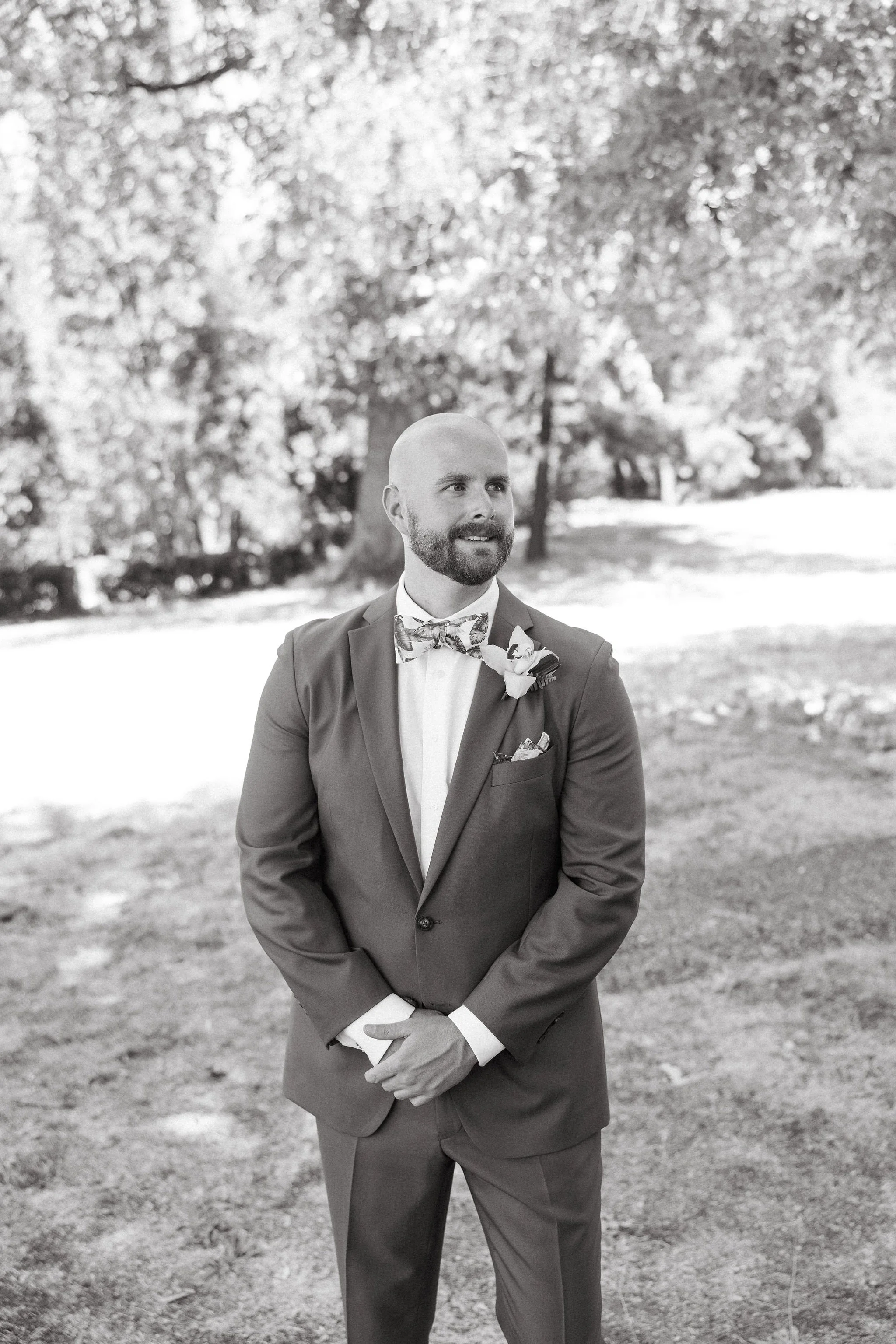 Man in a suit with a bow tie and boutonniere standing outdoors in a park with trees in the background.
