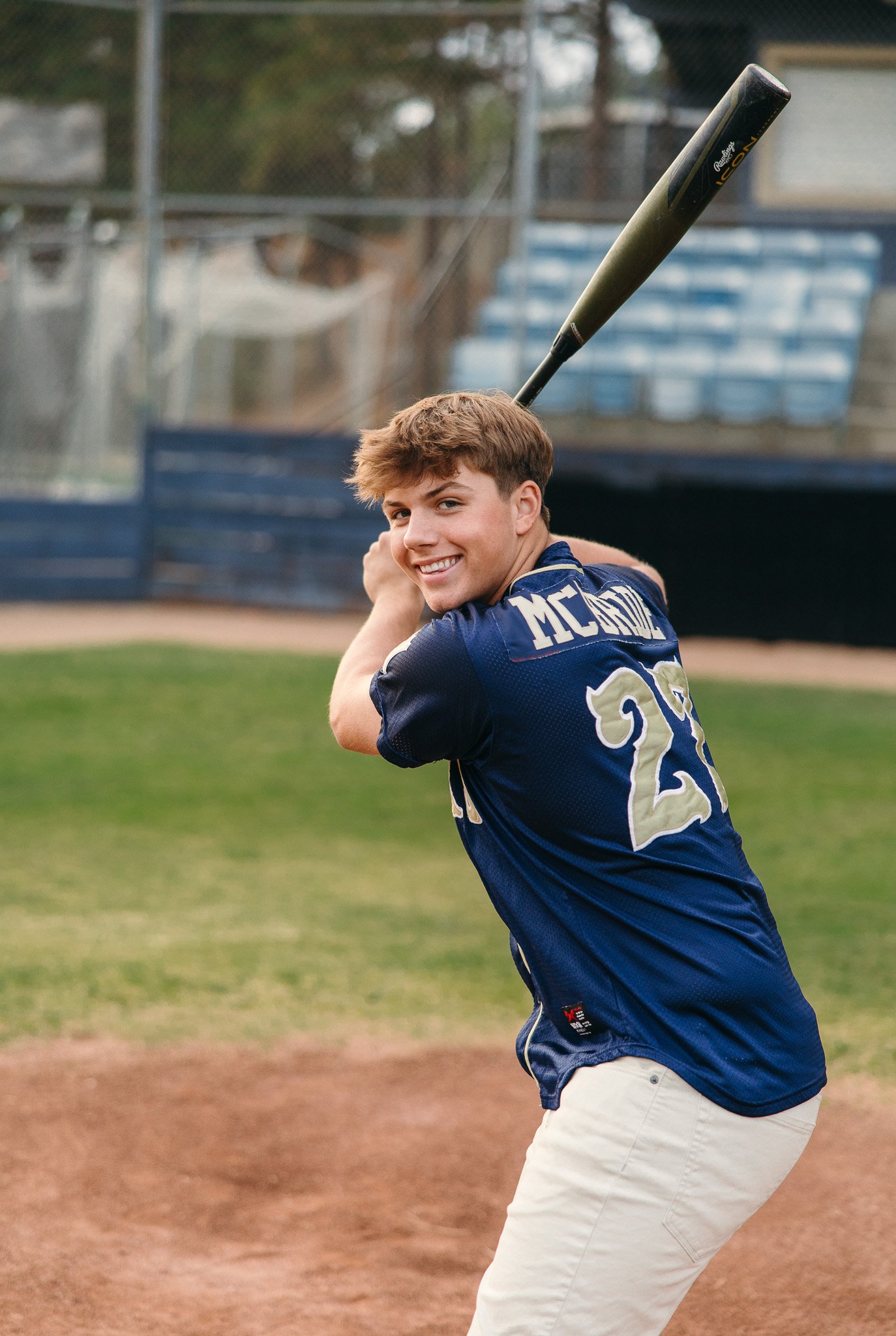 A young man in a navy blue baseball jersey and white pants, holding a baseball bat over his shoulder, smiling on a baseball field.