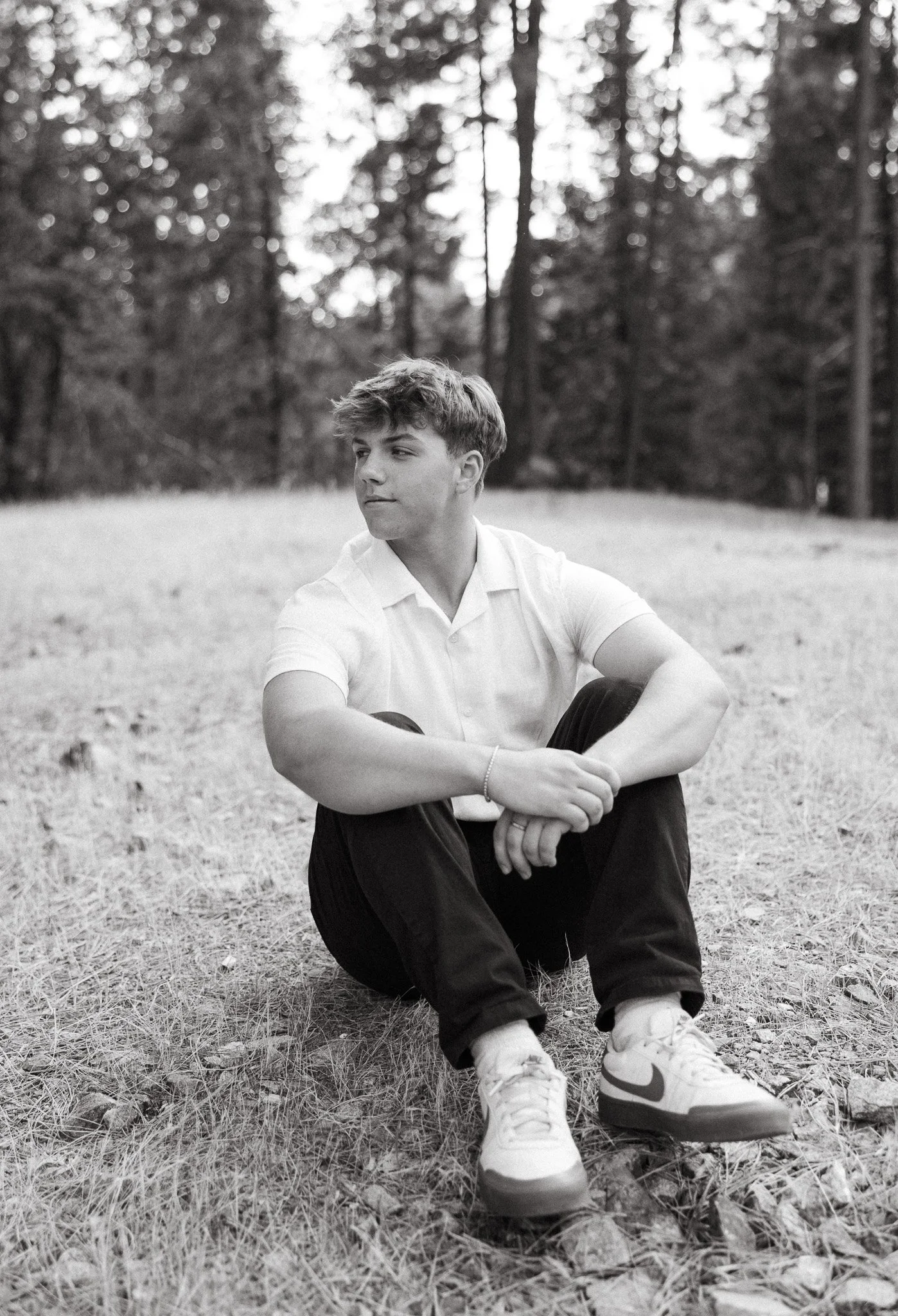 A young man sitting on the grass in a forested area, wearing a white polo shirt, dark pants, and white sneakers, looking to the side with a calm expression.