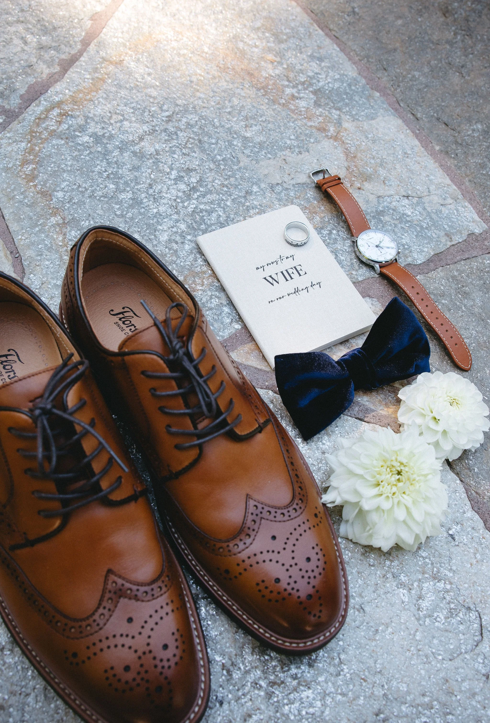 Brown leather dress shoes with black laces, a white wedding vow book titled 'My promise to my wife on our wedding day,' a black velvet bow tie, a gold watch with a brown strap, a silver ring, a silver ring, and white flowers, all arranged on a stone 