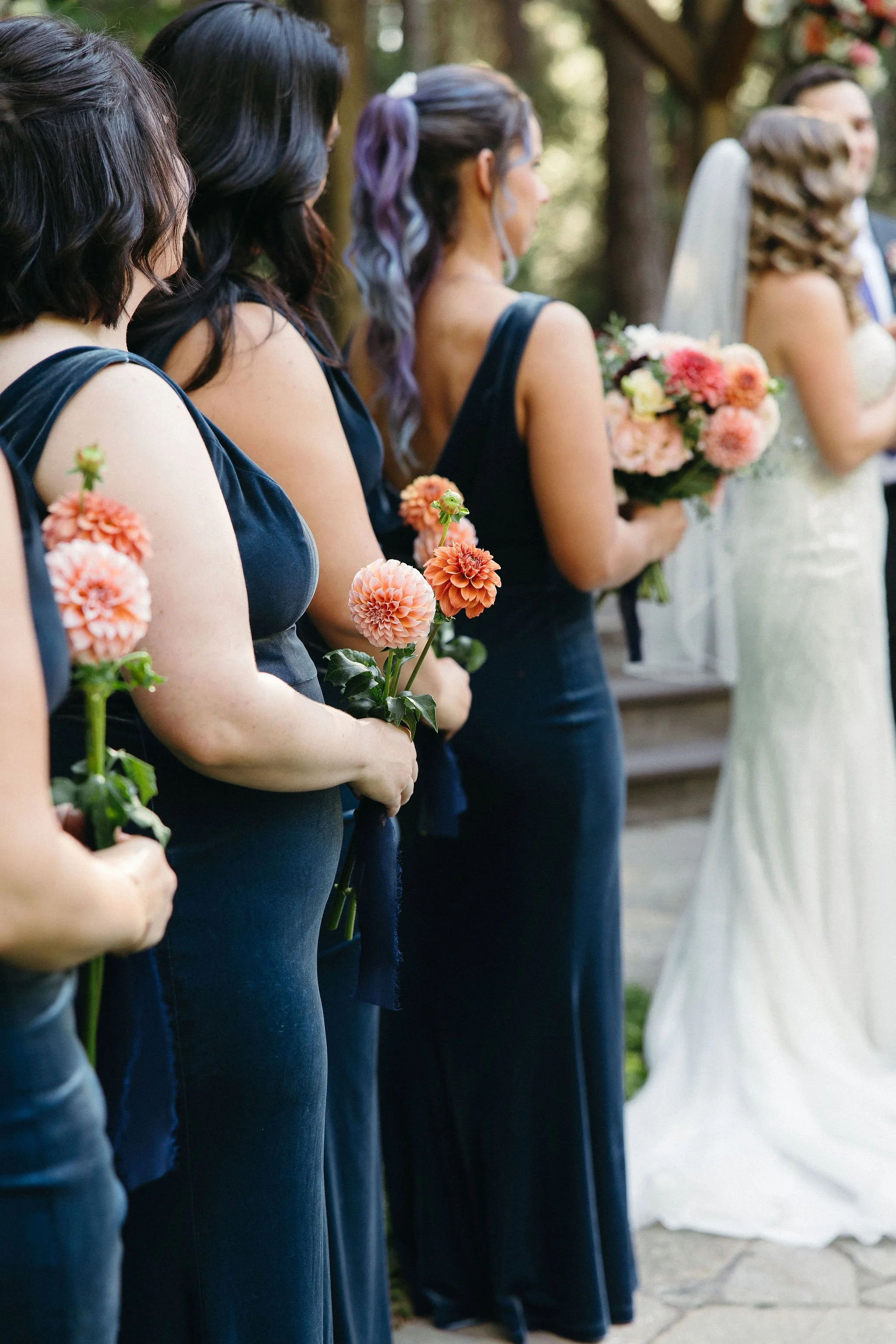 Line of women in navy dresses holding peach and pink flowers at a wedding ceremony outdoors.
