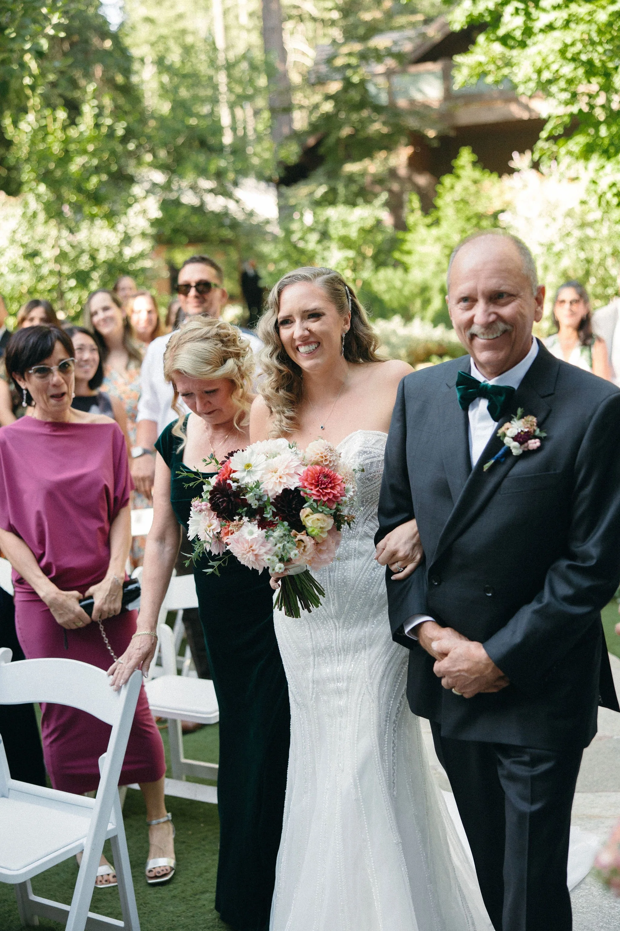 A bride in a white wedding dress holding a bouquet of pink, white, and purple flowers walking down the aisle with an older man in a suit. Guests are smiling and watching in the background outdoors amid greenery.