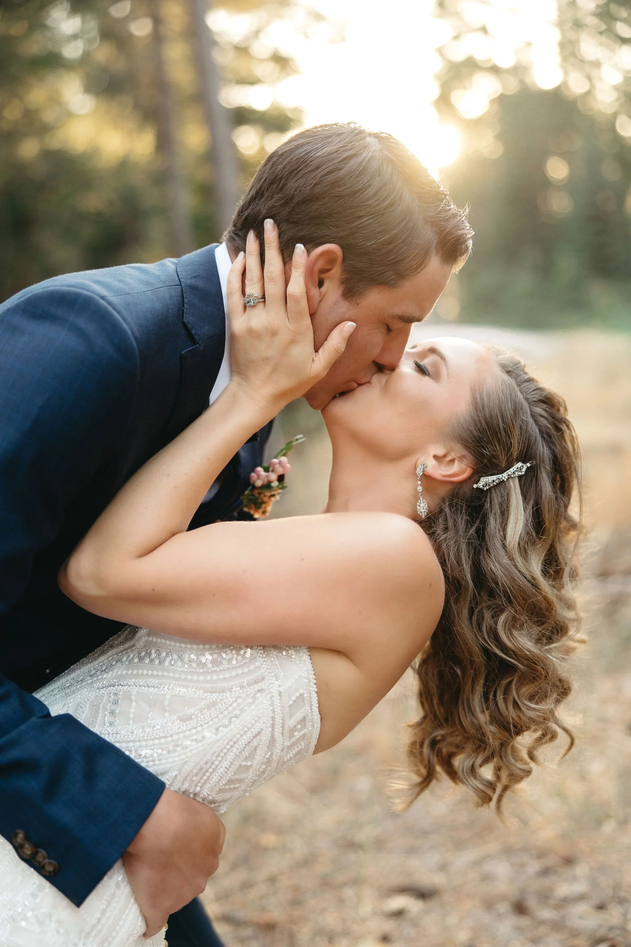 A bride and groom sharing a kiss outdoors during sunset, the groom leaning over the bride, who is holding his face with her hands, surrounded by trees.