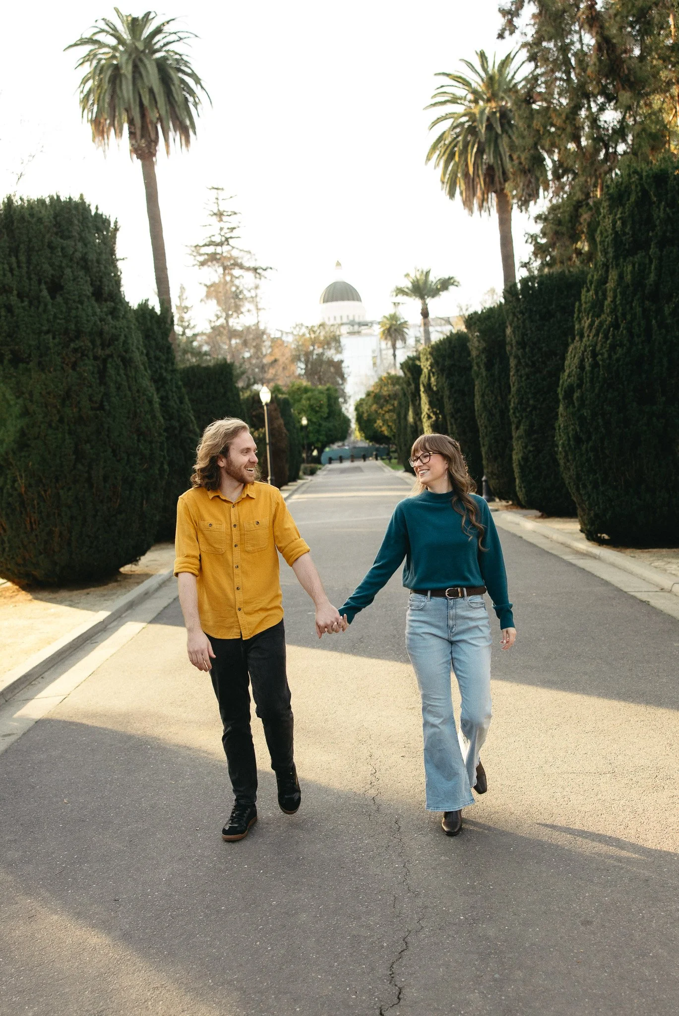 A young man and woman walk hand-in-hand down a paved street lined with tall, green hedges and palm trees. They are smiling and looking at each other. In the background, a dome-shaped building is visible, illuminated by soft sunlight.