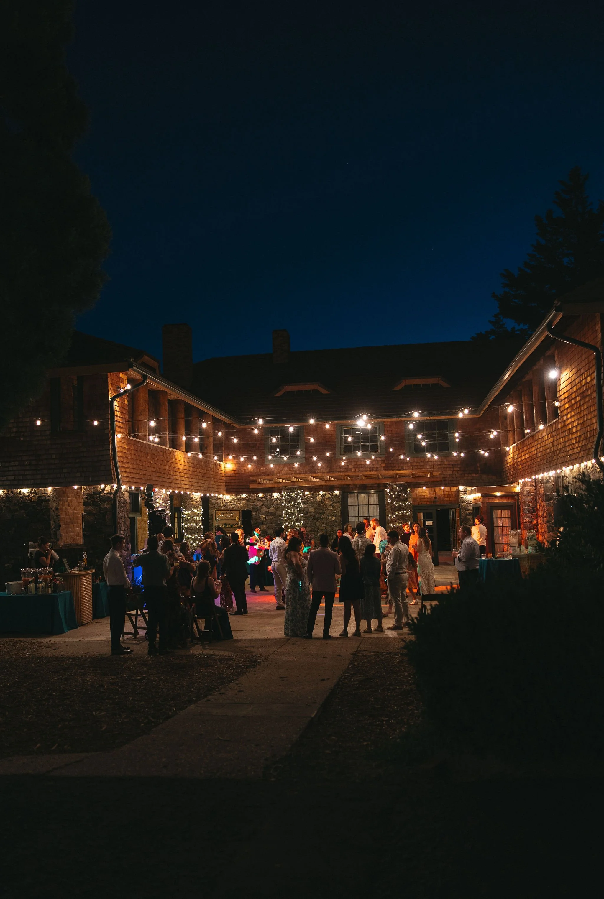 A nighttime outdoor party with string lights hanging from the walls of a large brick and stone building. People are mingling and dancing in the courtyard with tables on the sides.