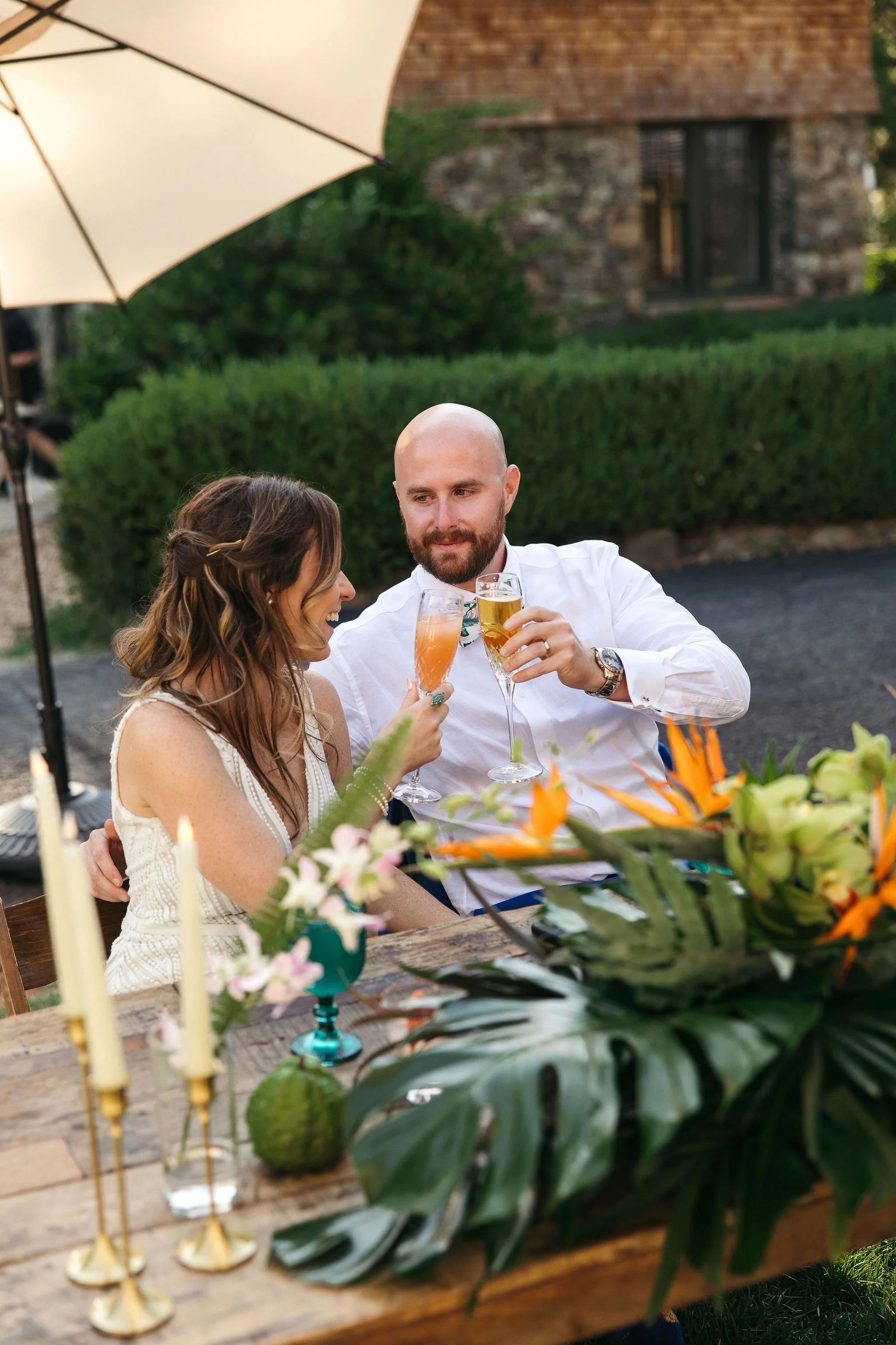 A couple at an outdoor party raising glasses for a toast, surrounded by floral arrangements and table decorations.