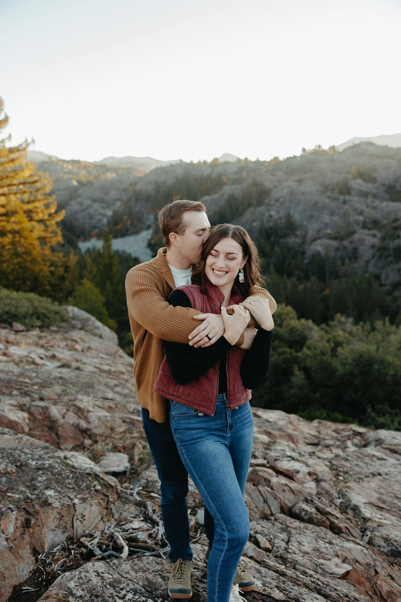 A couple standing outdoors on a rocky terrain, surrounded by mountains and trees, hugging each other and smiling with a clear sky in the background.