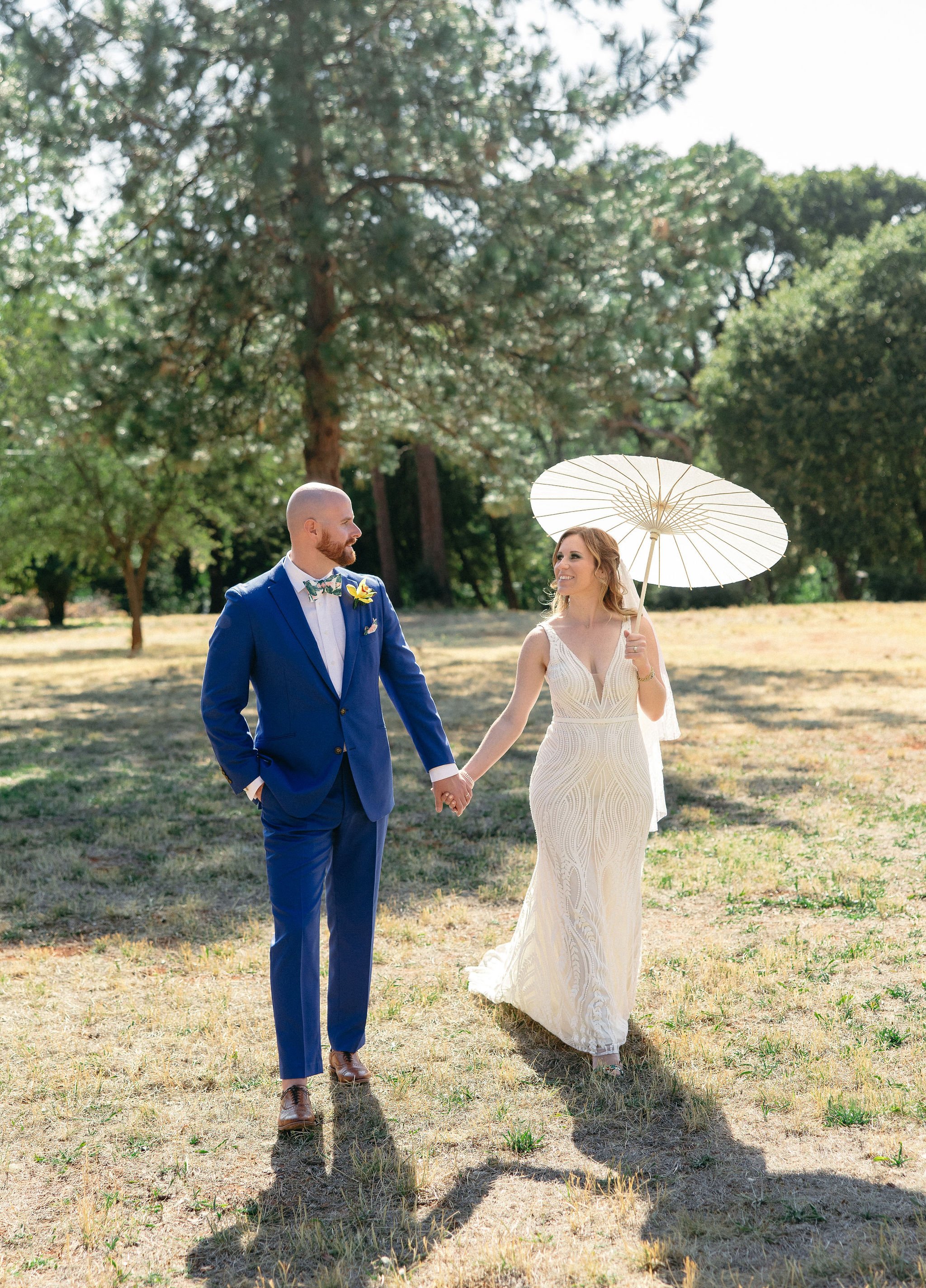 A bride and groom holding hands in an outdoor park setting, with the bride holding a white parasol and wearing a white wedding dress, and the groom in a blue suit with a bow tie.